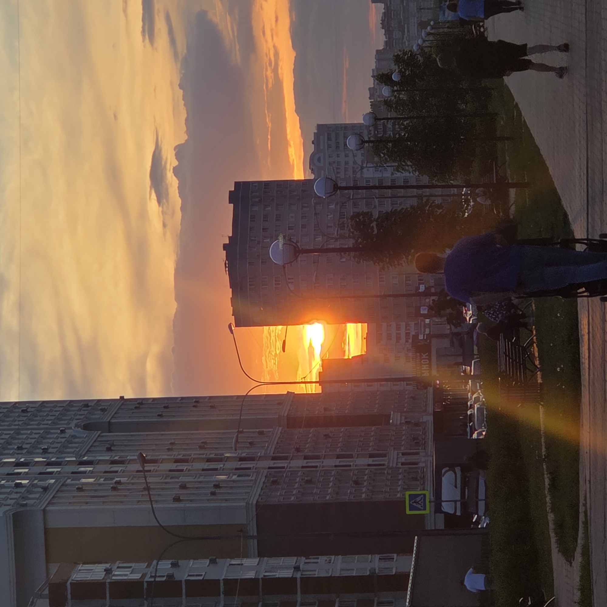 The image captures an urban street scene bathed in the warm glow of sunset. The sky is a dramatic canvas of orange and yellow, with scattered clouds highlighted by the setting sun. The sun itself is a brilliant, intense orb, positioned low on the horizon and visible between two large, multi-story city buildings, creating a powerful lens flare that stretches across the scene.
Two prominent tall buildings frame the left and center of the image, characterized by numerous windows and a repeating architectural pattern. A paved sidewalk or pedestrian path runs along the right side of the frame, bordering a strip of green grass. Several modern streetlights with spherical lamps are regularly spaced along the path. Sparse trees are visible in the grassy areas, their silhouettes dark against the bright sky.
In the mid-ground, a person wearing a blue shirt and dark pants is seen pushing what appears to be a stroller or cart along the sidewalk. Another individual in dark attire walks further ahead. Additional people are visible in the distance, though their features are indistinct.
Notable details include a blue and yellow pedestrian crossing sign on the left side of the path. Partial text, "ЫНОК", written in Cyrillic script, is visible on one of the buildings, suggesting a location like a "market" (РЫНОК). The overall impression is one of a peaceful evening in a city, with the vibrant sunset casting a serene atmosphere over the urban landscape.