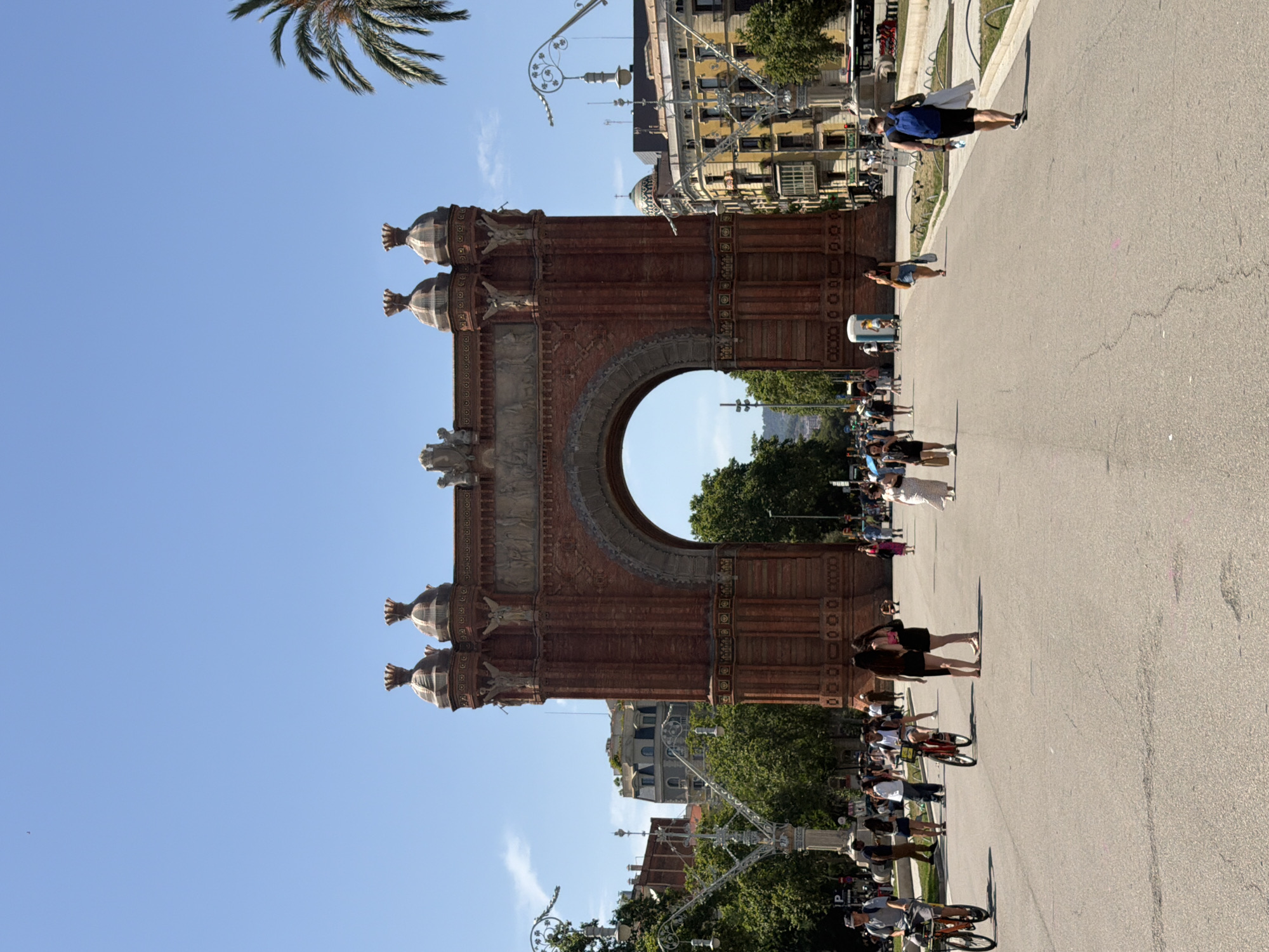 This is a vertical, eye-level, outdoor, daytime photograph of the Arc de Triomf in Barcelona, Spain. The massive, reddish-brown brick arch is the central and dominant feature of the image, spanning the width of the frame. It is ornately decorated with carvings and architectural details. The arch is situated at the end of a wide, paved pathway that extends into the distance, lined with trees and lampposts. The pathway is populated by numerous people, some walking, some cycling, and some standing in groups. The sky is a clear, bright blue with a few wispy clouds. The architecture of the surrounding buildings, visible beyond the arch and along the pathway, is characteristic of Barcelona, with balconies and varied rooflines. A palm tree is visible on the right side of the frame.