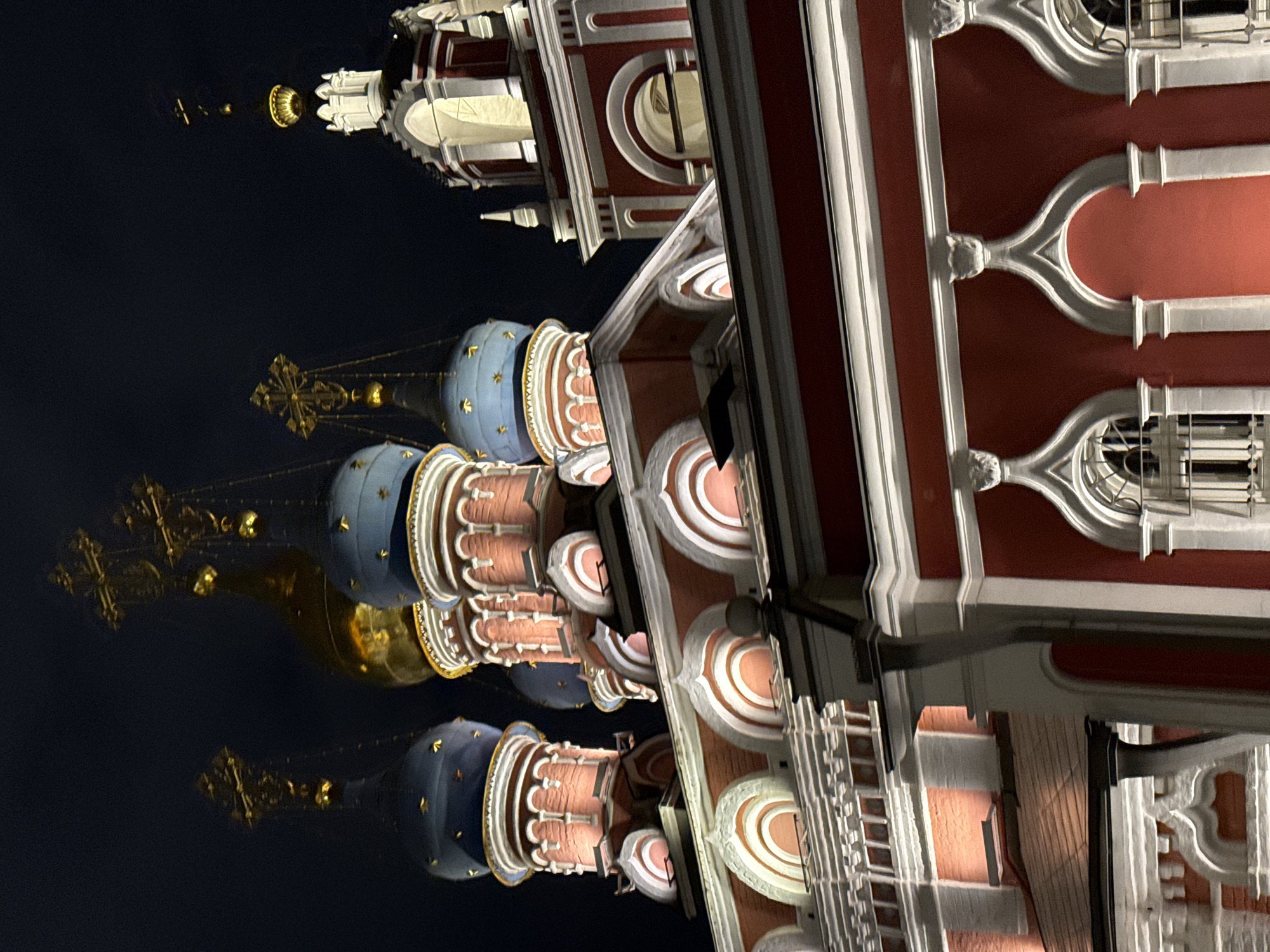 The image shows a nighttime view of the domes and architecture of St. Basil's Cathedral in Moscow, Russia. The domes are brightly lit, showcasing their colorful details, including blue domes with gold stars and onion-shaped domes with intricate designs. The architectural elements of the cathedral include red walls with white decorative elements. The sky in the background is dark, contrasting with the illuminated building. The perspective is from a low angle, looking upwards.