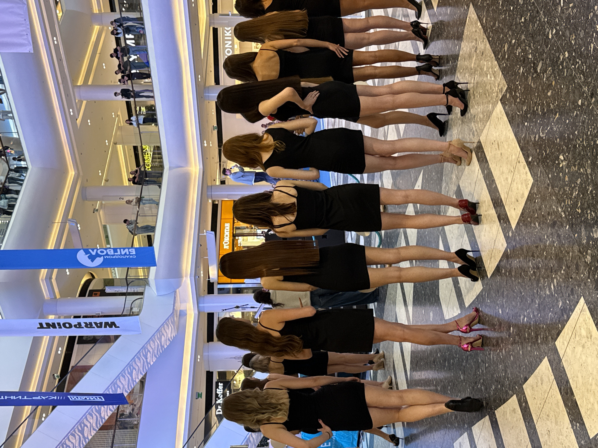 The image shows the interior of a modern shopping mall in Moscow, Russia, likely in the evening given the lighting. A group of at least nine young women, all wearing short, sleeveless black dresses and high heels, are standing in a line facing away from the camera. They appear to be participating in an event or performance. Some are standing close together, while others are spaced slightly apart. In the background, escalators, railings, and various shop signs are visible. A few people are seen on the upper levels of the mall, observing the scene below. The floor is tiled with a geometric pattern of light and dark squares. The overall atmosphere suggests a public event or gathering within a commercial space.