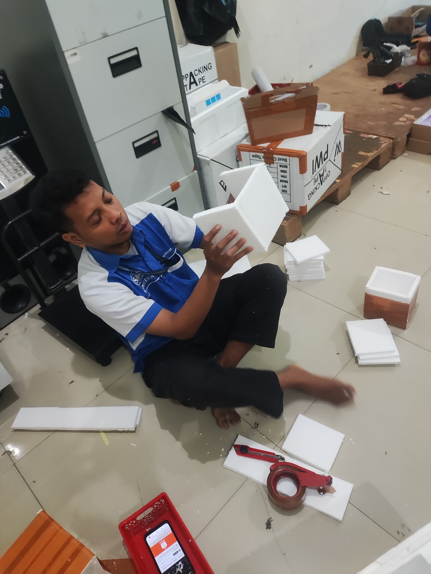 The image depicts a man sitting on the floor in a room that appears to be a warehouse or a workshop, likely in Pepelegi, Indonesia. He is wearing a blue and white polo shirt and black pants, and he is in the process of assembling white styrofoam packaging. Various pieces of styrofoam, some assembled into boxes, are scattered around him on the tiled floor. In the background, there are filing cabinets, stacked boxes, and a wooden pallet. A red utility knife and a roll of tape are also visible on the floor. The lighting suggests it might be daytime, possibly with artificial lighting supplementing natural light. The overall scene conveys an atmosphere of a production or packing environment.