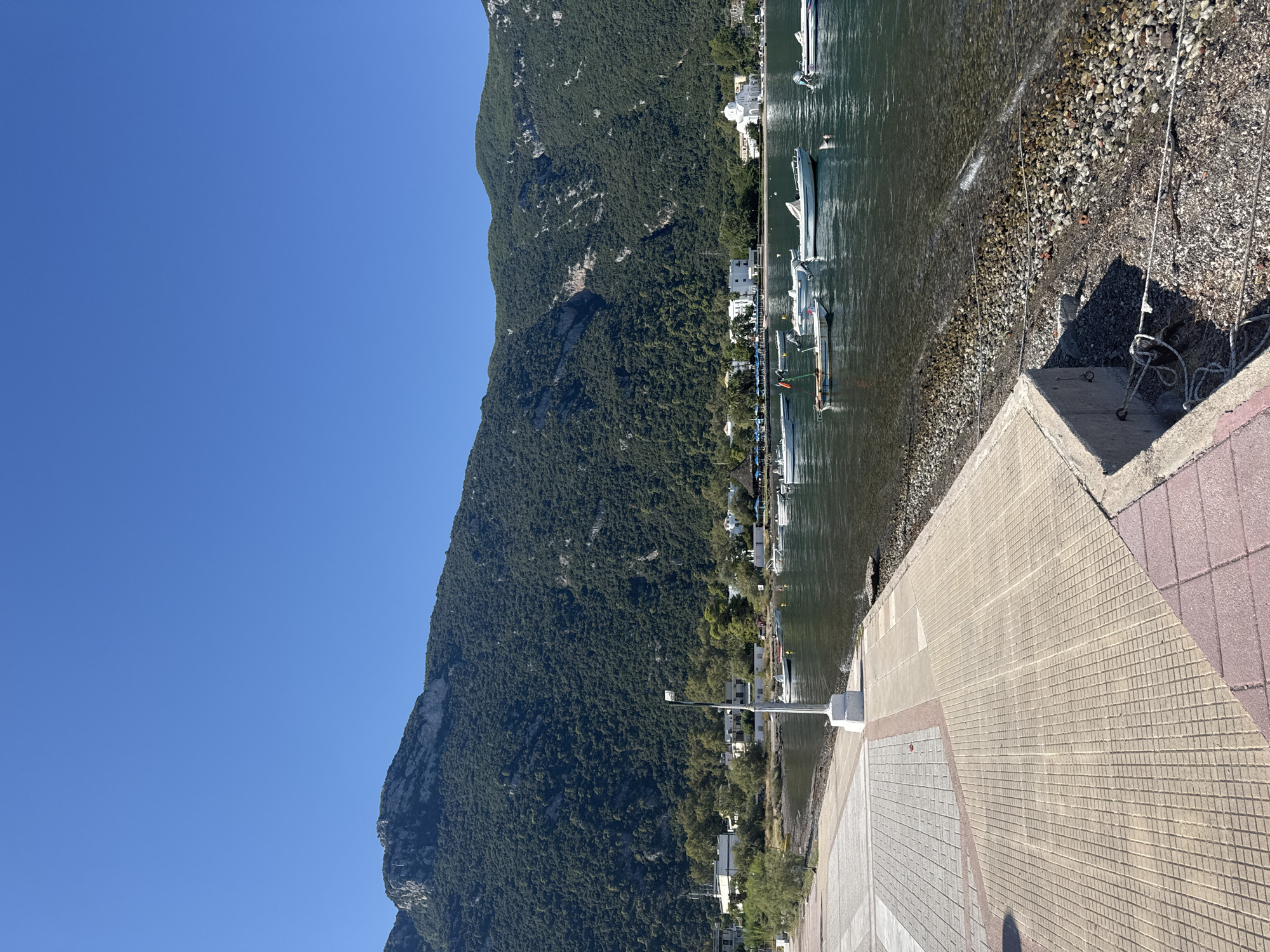 This is a photograph taken on a sunny day, depicting a coastal scene in the Municipal Unit of Kamena Vourla, Greece. The foreground features a paved walkway with a patterned surface of interlocking tiles. Adjacent to the walkway is a stony shoreline leading into the water. Several small boats are moored in the clear, calm water.
In the midground, a cluster of white buildings, some appearing to be houses and others a church with a distinctive dome, are visible along the coastline. Trees and vegetation surround these structures.
Dominating the background is a large, verdant mountain covered in dense green foliage. The mountain rises steeply from the coastline, its slopes intricately textured by the vegetation. The sky above is a clear, unblemished blue. A lone lamppost stands on the edge of the walkway, facing the sea. The overall impression is one of a tranquil, sun-drenched coastal village nestled at the foot of a substantial mountain.