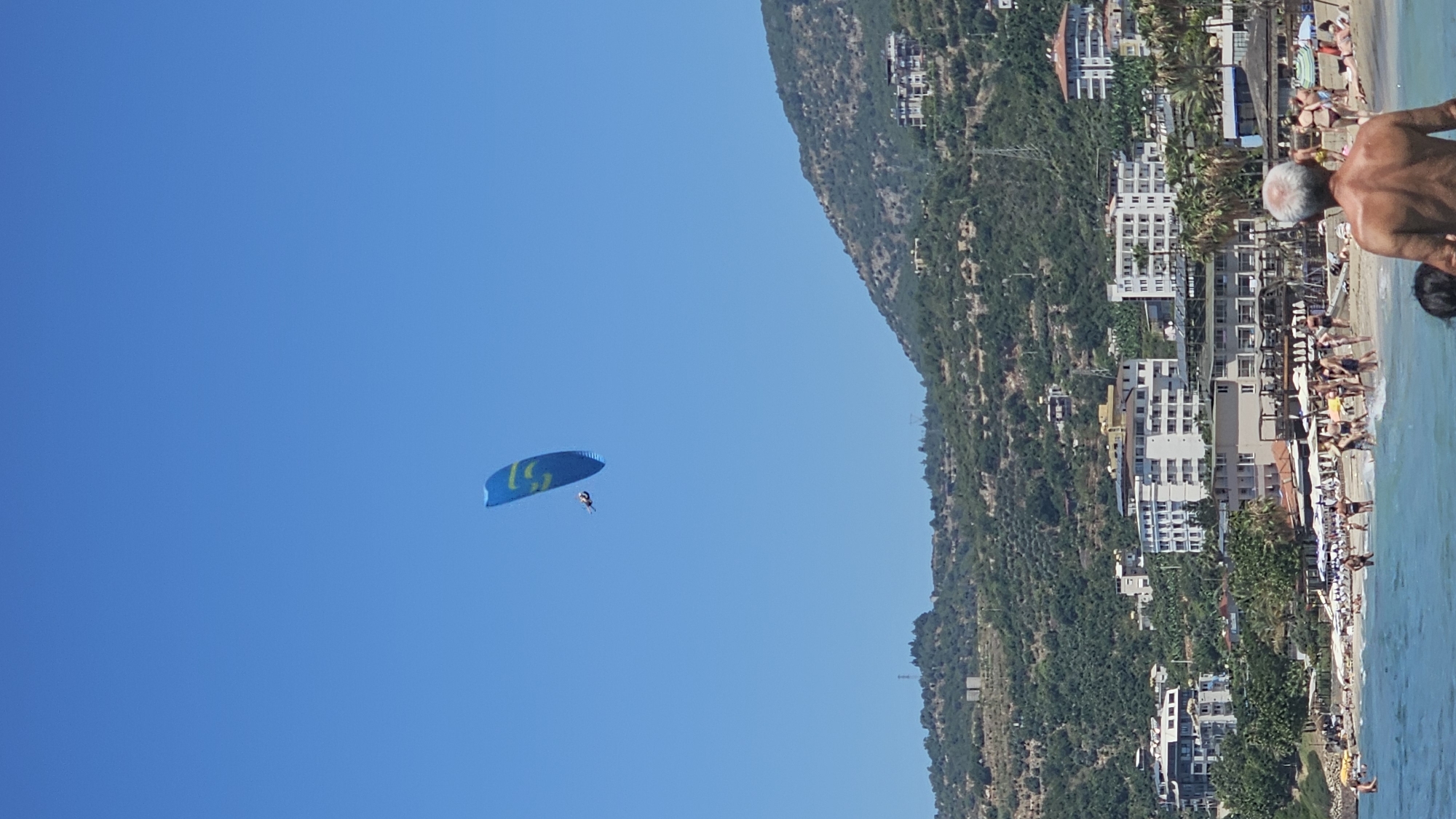 In Alanya, Turkey, a bright blue paraglider with yellow markings soars through a clear blue sky. The paraglider is carrying a person, and it is positioned high above a scenic coastal town. The town is built on the slopes of a lush green mountain, with numerous white buildings nestled amongst the trees. Below the mountain, a crowded beach is visible, with many people enjoying the sun and the sea. Beach umbrellas dot the sand, providing shade. The water at the edge of the beach is a vibrant turquoise, with gentle waves lapping at the shore.