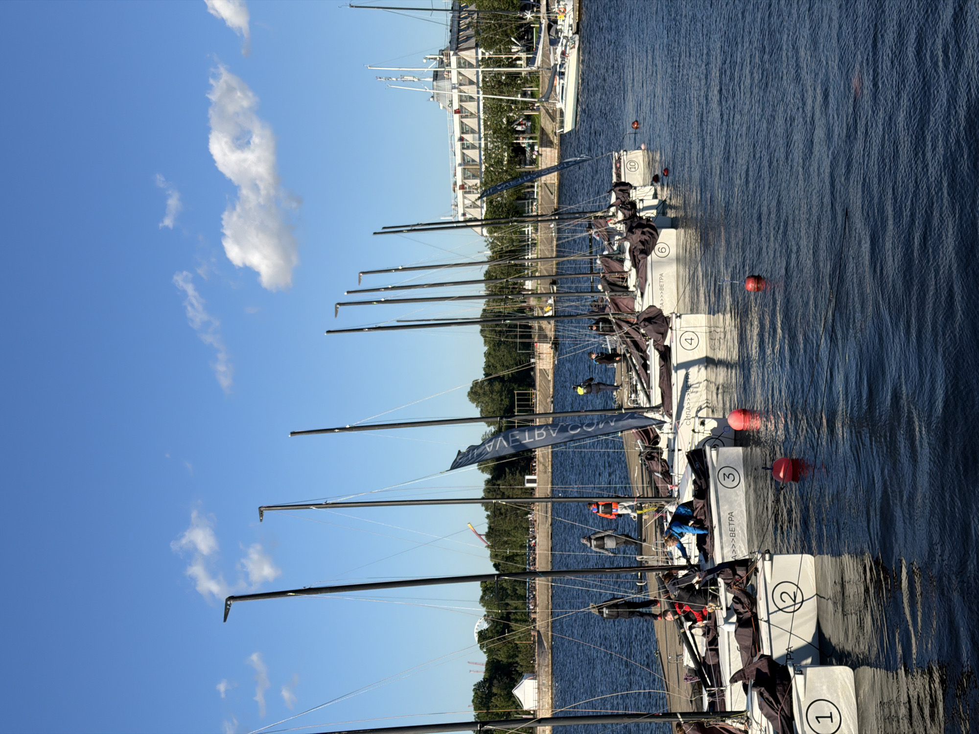 The image shows a fleet of sailboats docked in a marina. The boats are lined up in parallel, with their masts reaching towards the blue sky. Several people are visible on the decks of the boats, some tending to equipment and others observing the surroundings. The water is calm, reflecting the blue sky and the white hulls of the boats. In the background, a modern building with multiple windows and trees can be seen along the waterfront, suggesting an urban setting. The scene appears to be taking place on a sunny day, likely in the late morning or early afternoon given the bright light and clear skies. Red buoys are floating in the water near the boats. Some of the boats have numbers and text printed on their hulls. Visible text includes "1", "2", "3", "4", "6", and "10", as well as "А>>>ВЕТРА" and "СИЛА>>>ВЕ". The overall impression is one of a regatta or a sailing event in progress.