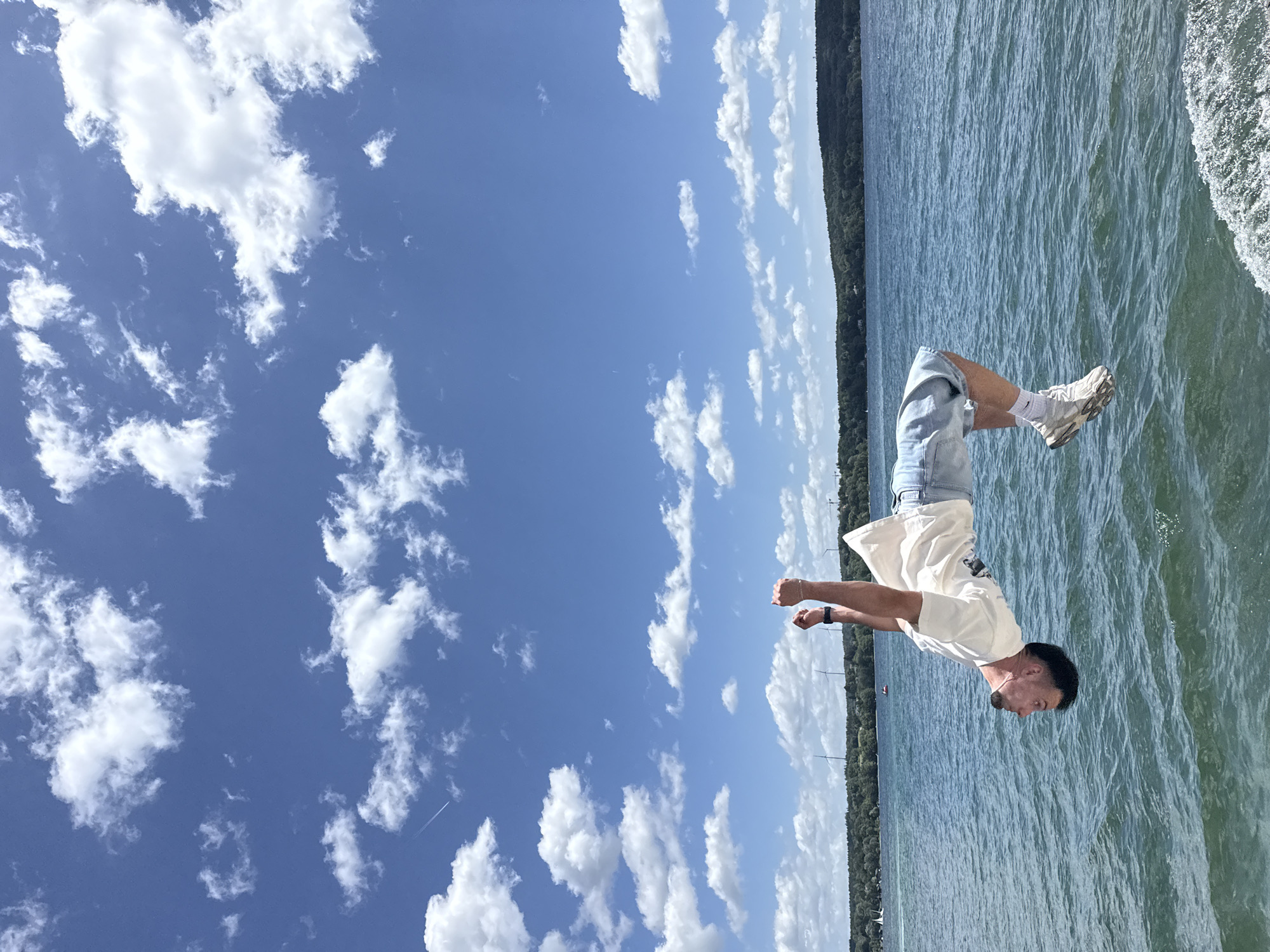 A man is captured mid-air, performing a backflip over the water of Lake Starnberg, Germany. He is wearing a white t-shirt, denim shorts, white socks, and athletic shoes. His body is inverted, with his head pointing downwards and his arms raised above him, clenched into fists. The lake is calm with small ripples, reflecting the clear blue sky dotted with white clouds. In the background, a tree-lined shore stretches across the horizon, with a few wind turbines visible. The overall scene depicts an athletic feat against a scenic natural backdrop.