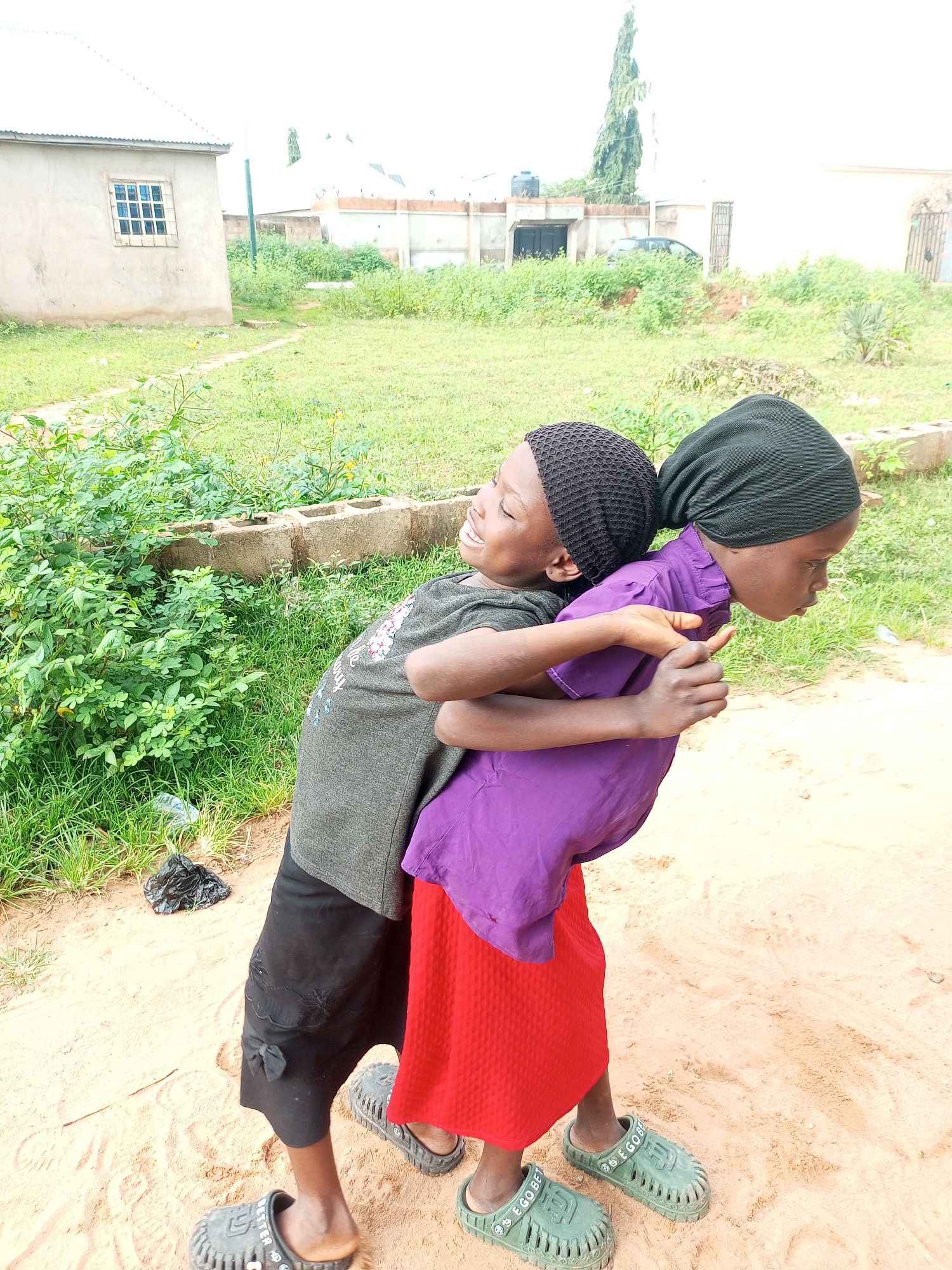 Two children are interacting in an outdoor environment that appears to be a sandy, unpaved area with grass and some low-lying vegetation. The child in front, facing to the right of the frame, is wearing a black head covering, a grey t-shirt with a floral design, black shorts with a bow detail, and black clogs. This child has their eyes closed and is laughing. The child behind, facing to the left, is wearing a black head covering and a purple top with a red skirt, and green clogs. This child has their arms around the other child's waist. In the background, there is a light-colored building with a metal roof and a small window, a chain-link fence, and more vegetation. The ground is a mixture of sand and dirt. The overall lighting suggests it is daytime. The scene is likely a residential area or a public outdoor space in Tempure, Nigeria.