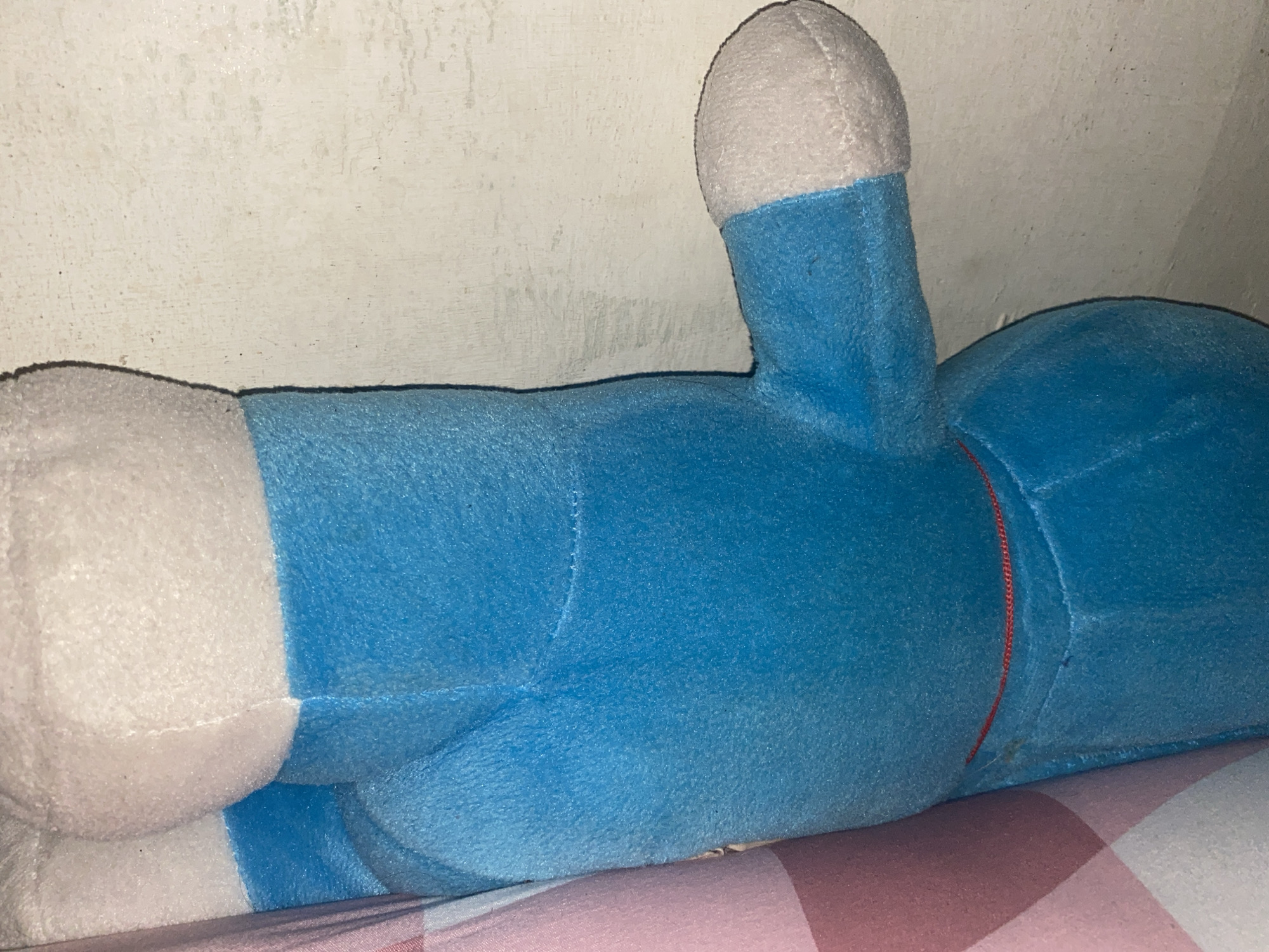 A blue plush toy is lying on a pink and white checkered surface, possibly a bed or a blanket. The toy appears to be a character from a cartoon or anime, with a round white head and a blue body. One of its arms is raised, and it has white accents on its hands and feet. The background is a textured, off-white wall. The lighting is dim, suggesting the photo was taken indoors, possibly at night or in a room with limited light. There is no discernible text in the image. The overall impression is one of a child's toy placed in a resting position.