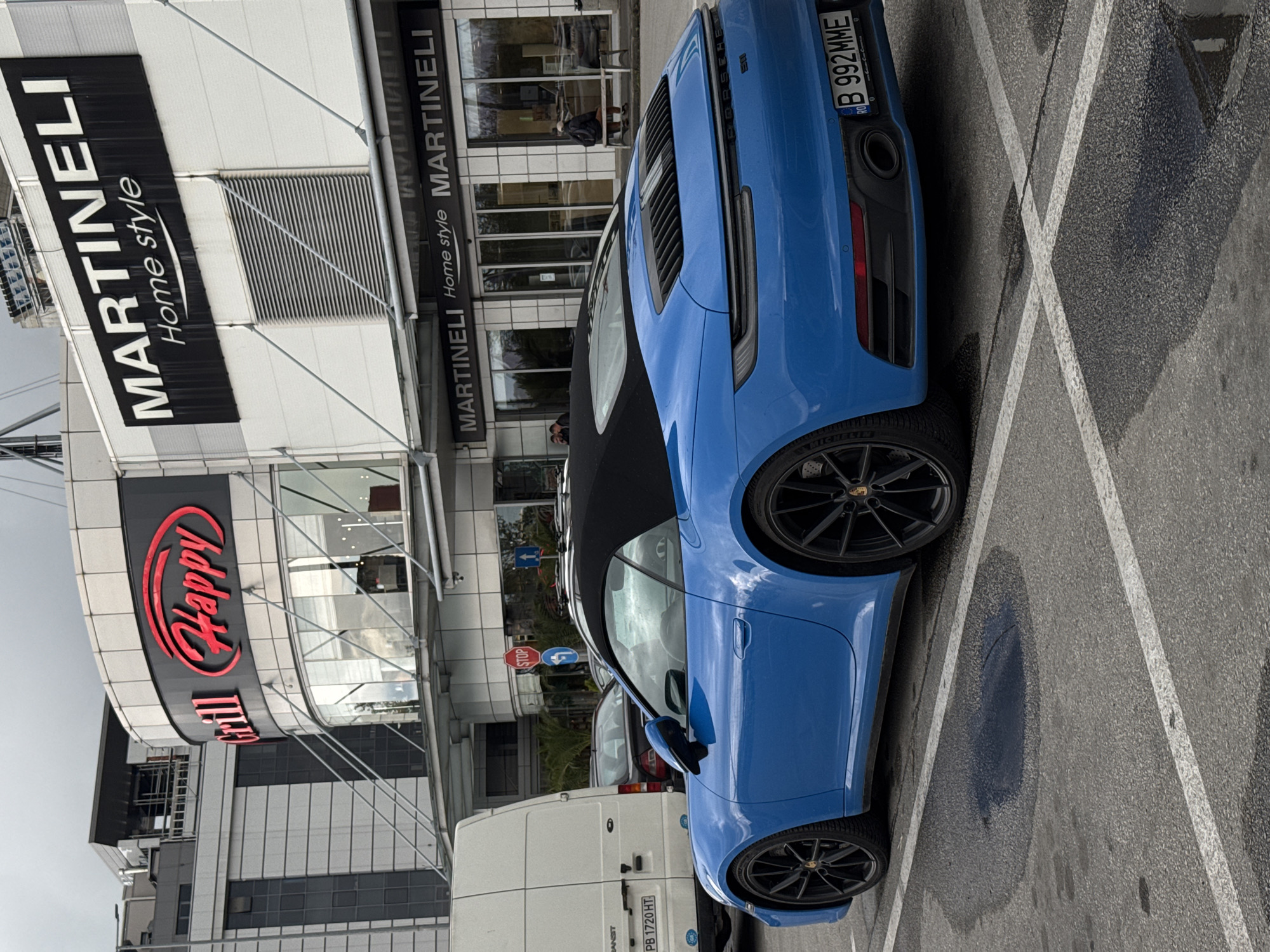 This outdoor image displays a vibrant blue Porsche 911 convertible parked in an asphalt lot. The car, viewed from its rear-right side, features a black soft-top, black multi-spoke wheels with Michelin tires, and a visible Romanian license plate "B 992MME." The word "PORSCHE" and "911" are subtly visible on the rear of the car.
In the background stands a modern commercial building with prominent signage. A large banner reads "MARTINELI Home style" in white on black, with the name "MARTINELI" repeated elsewhere on the facade. Below this, a red oval logo marks "Happy Grill." A few people can be seen near the entrance of the "MARTINELI" store.
To the left of the Porsche, a white Ford Transit van is parked, bearing a Bulgarian license plate "PB 1720 HT," confirming the location context of Plovdiv, Bulgaria. Nearby, a "STOP" sign and blue directional arrow signs are mounted on a pole.
The parking lot surface is marked with white lines and shows several puddles, suggesting recent rainfall. The sky is overcast, indicating a daytime scene, likely under cloudy weather conditions. The overall setting is a typical commercial area or shopping center parking lot.