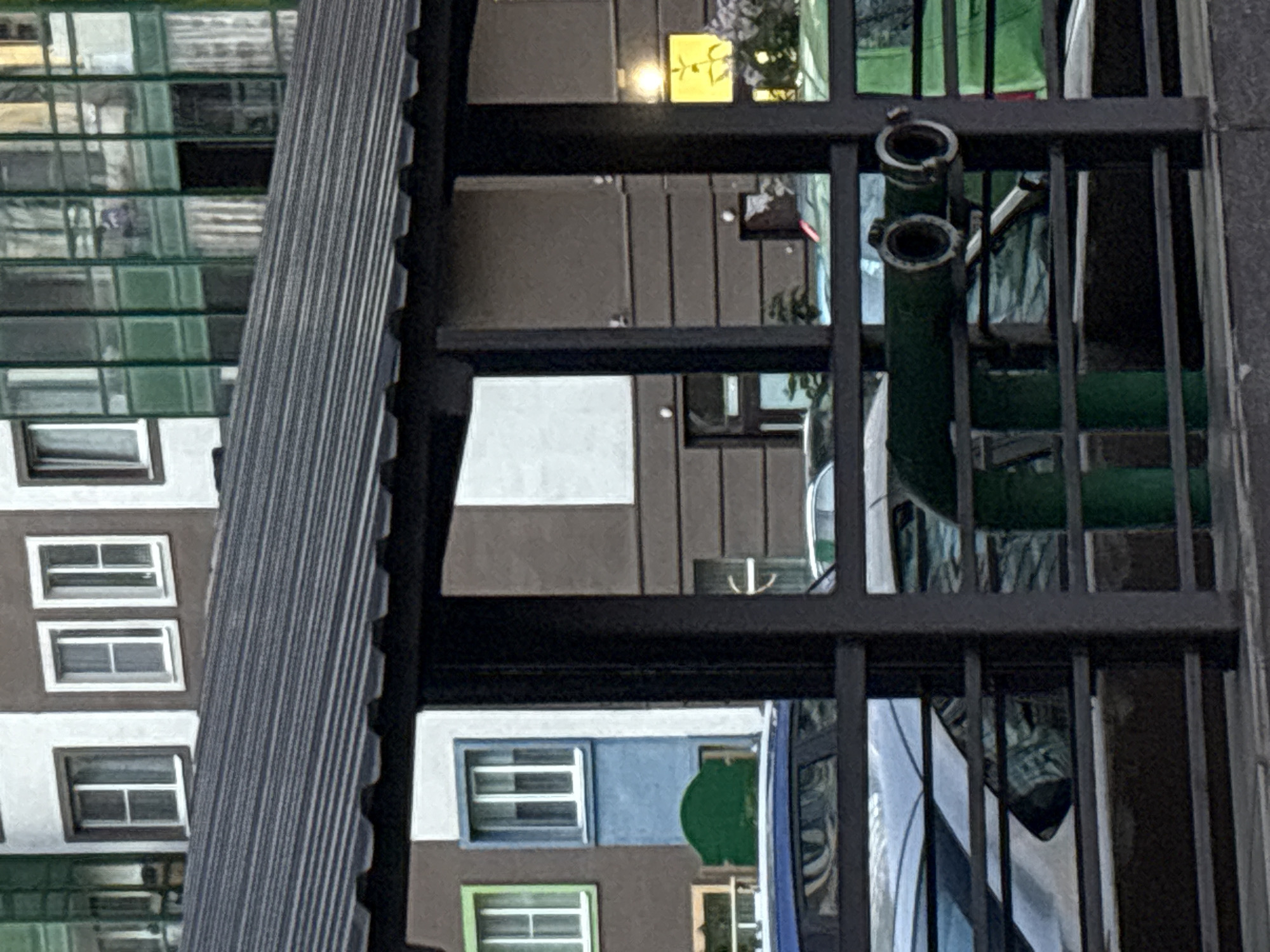 This is a medium-shot, eye-level, outdoor photograph of a modern building facade with a covered walkway in the foreground. The building is multi-story with a combination of white, brown, and green sections. Windows vary in style and placement, with some appearing to be modern glass panels and others more traditional. The covered walkway has a dark, corrugated metal roof supported by dark metal posts. Behind the walkway, cars are parked, and a green pipe with two circular openings is visible. The overall scene suggests an urban environment, possibly a residential or commercial area. The location is identified as Unknown, Russia.