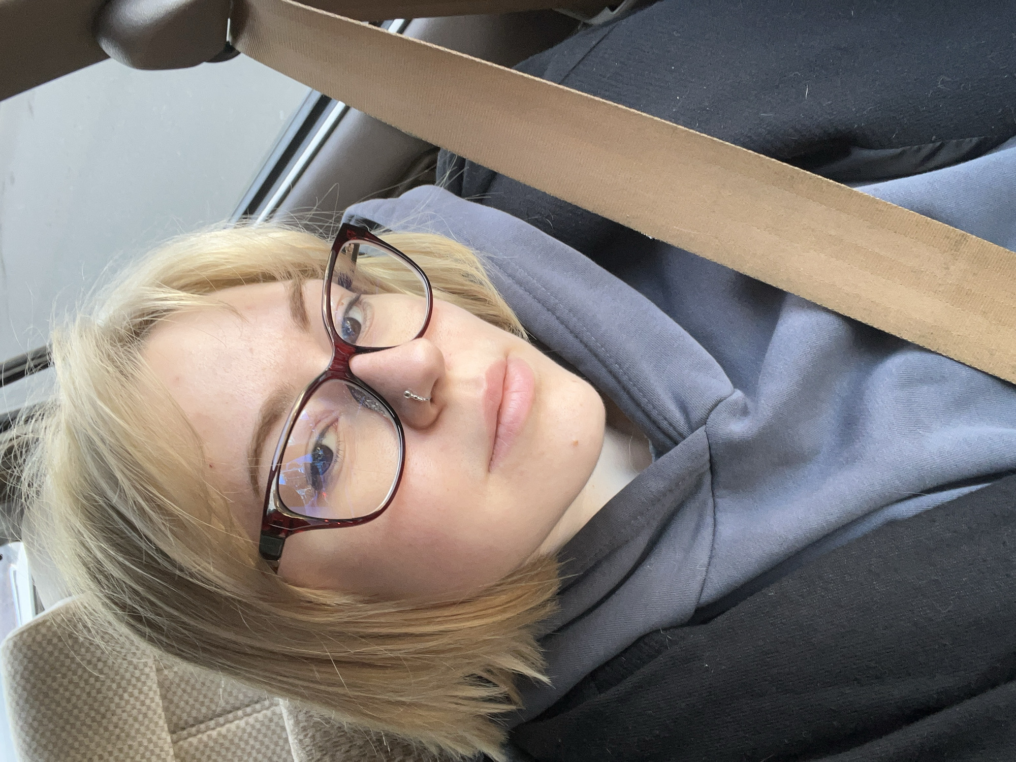 A fair-skinned person with blond hair, wearing glasses and a nose ring, is positioned in what appears to be a car. The person is wearing a gray hooded sweatshirt and a black jacket. A tan seatbelt is across their body. The interior of the car includes beige upholstery. The specific location in Odesa, Ukraine cannot be determined from the image.