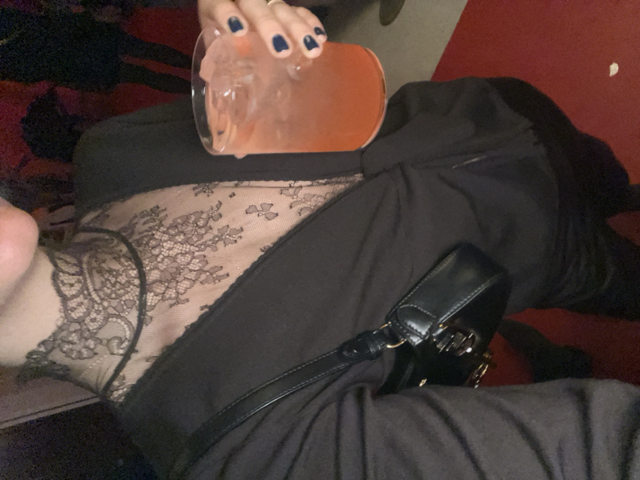 A person in black clothing is holding a drink with ice in a glass. The top appears to be a sheer lace with a solid black trim. A small black purse sits on the person's lap. The background features a red surface. The person's hand is visible, adorned with a ring and dark blue nail polish.