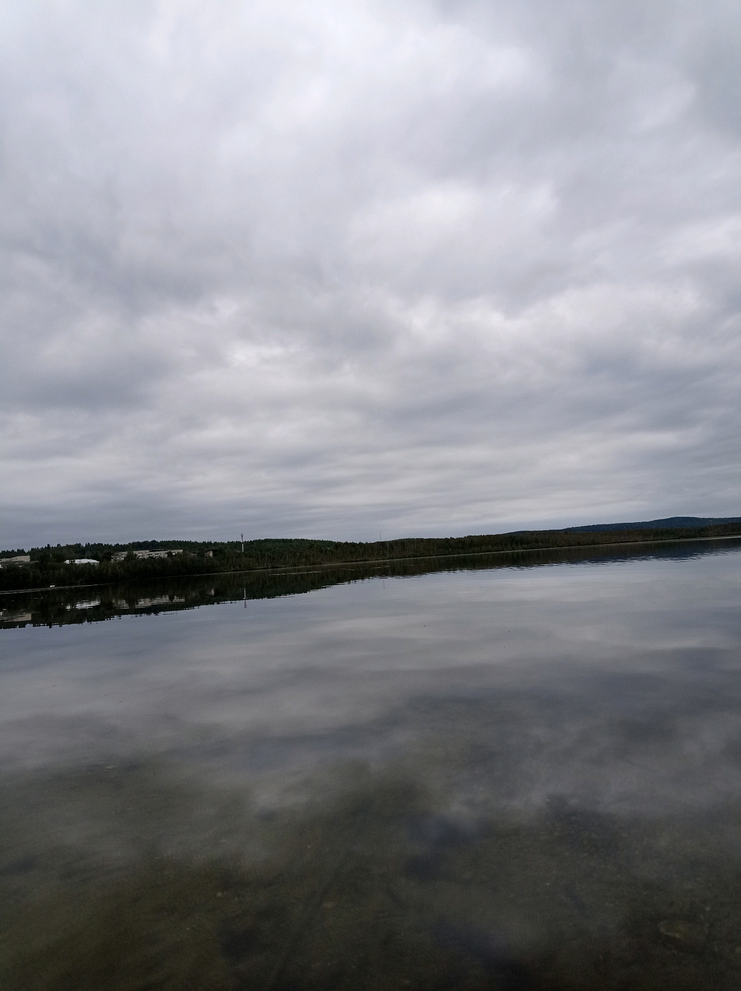 The image shows a serene, overcast day at a body of water, likely a lake or a wide river, in Degtyarsk, Russia. The sky is filled with dense, gray clouds, creating a diffused light. The water's surface is calm, reflecting the cloudy sky and the treeline of the distant shore. The far shore is lined with dark green trees, interspersed with some buildings and structures that appear to be part of a settlement. A tall, slender tower, possibly a communication tower, is visible among the trees on the right side of the frame. The foreground shows the shallow bottom of the water, with visible pebbles and sand. There are no people or animals visible in the image. The overall atmosphere is one of quietude and natural beauty, characteristic of a northern landscape under a cloudy sky.