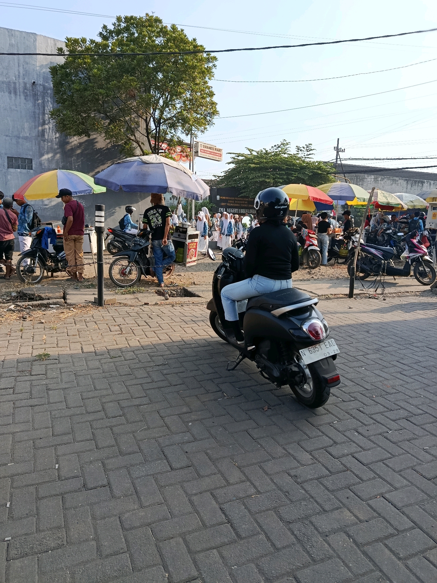The image depicts a street scene in front of SMA Negeri 1 Bululawang in Bululawang, Indonesia, on a sunny day. Several students in uniform are visible, some walking and some gathered under colorful umbrellas. A woman wearing a black helmet and casual clothing rides a black scooter in the foreground, facing away from the camera. Other individuals, likely vendors or parents, are present near food stalls set up under umbrellas. Motorcycles are parked along the roadside. The ground is paved with interlocking concrete blocks. A large tree shades a portion of the scene. The overall atmosphere suggests a busy area, possibly near a school during dismissal or a local market.