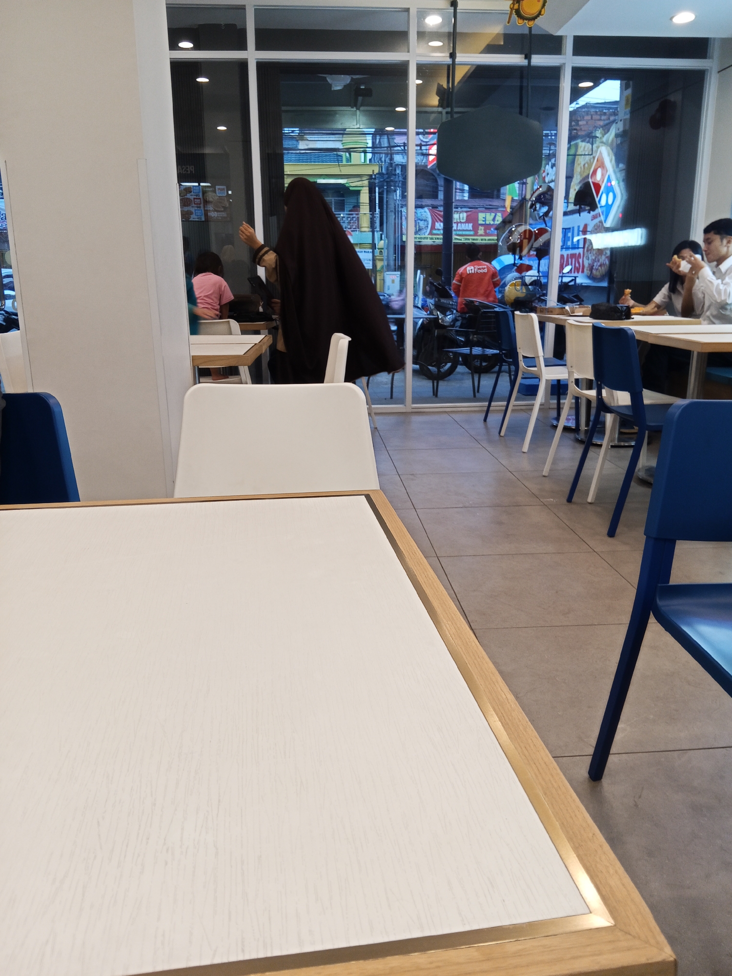 The image depicts the interior of a Domino's Pizza restaurant in Lubuklinggau, Indonesia. The foreground shows a white table with a wooden frame. In the middle ground, several chairs are visible, including blue chairs and white chairs. Through a large glass window, the exterior of the restaurant is visible, showing a street with buildings and motorbikes. Inside the restaurant, several patrons are present. A person wearing a dark headscarf and brown hijab is standing near the window, reaching towards a table. Further back, a person in a pink shirt is seated at a table. Two individuals, a woman and a man, are seated at a table in the background, with the woman holding a phone and the man holding food. The environment appears to be a typical fast-food dining area.