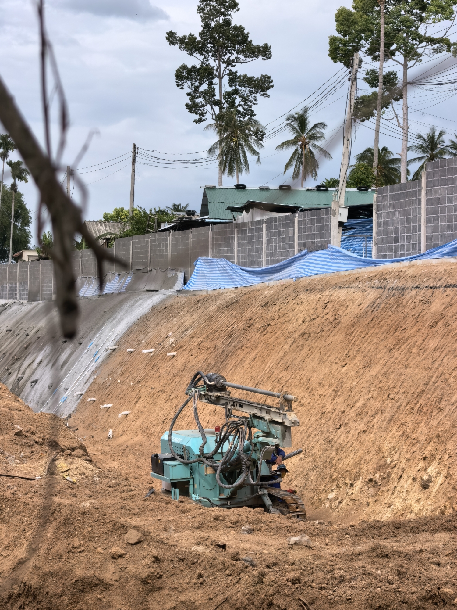 The image depicts a construction site in the Ko Samui District of Thailand. In the foreground, a teal-colored drilling rig is positioned on loose soil. A worker, wearing a blue uniform and a face mask, is operating the drill. Behind the rig, the ground slopes upwards, revealing exposed soil and a reinforced retaining wall. Sections of the retaining wall are covered with blue and white striped tarps. In the midground, a grey concrete block wall forms a barrier. Beyond the wall, several buildings with green roofs are visible. Tall trees, including palms and a large deciduous tree, dominate the background, along with overhead power lines and a cloudy sky. The scene suggests ongoing construction work, likely for soil stabilization or foundation preparation, in a tropical environment.