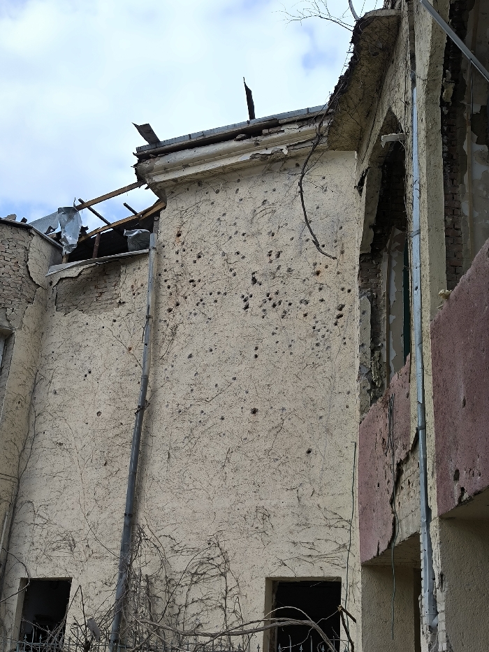 The image depicts a severely damaged building facade in Vinnytsia, Ukraine, under an overcast sky. The main wall, constructed of light beige stucco, is extensively pockmarked with numerous dark, irregular indentations, consistent with shrapnel or ballistic impacts. Significant cracking and peeling of the stucco are visible across the surface. A network of dry, bare vines is entangled on the wall, and a grey vertical downspout runs along its left side.
The building's roofline shows considerable structural damage, with exposed wooden rafters and crumpled metal sheeting indicating partial collapse. Portions of the decorative cornice below the roof are broken and displaced. To the right, an adjoining section of the building exhibits further destruction, including exposed brickwork, a partial window opening revealing a dark interior and a blue-green frame, and a peeling reddish-brown exterior finish. At the lower section of the main wall, two distinct dark, rectangular apertures, likely former windows or doorways, are visible amidst a tangle of bare branches. No individuals or active subjects are present in the scene.