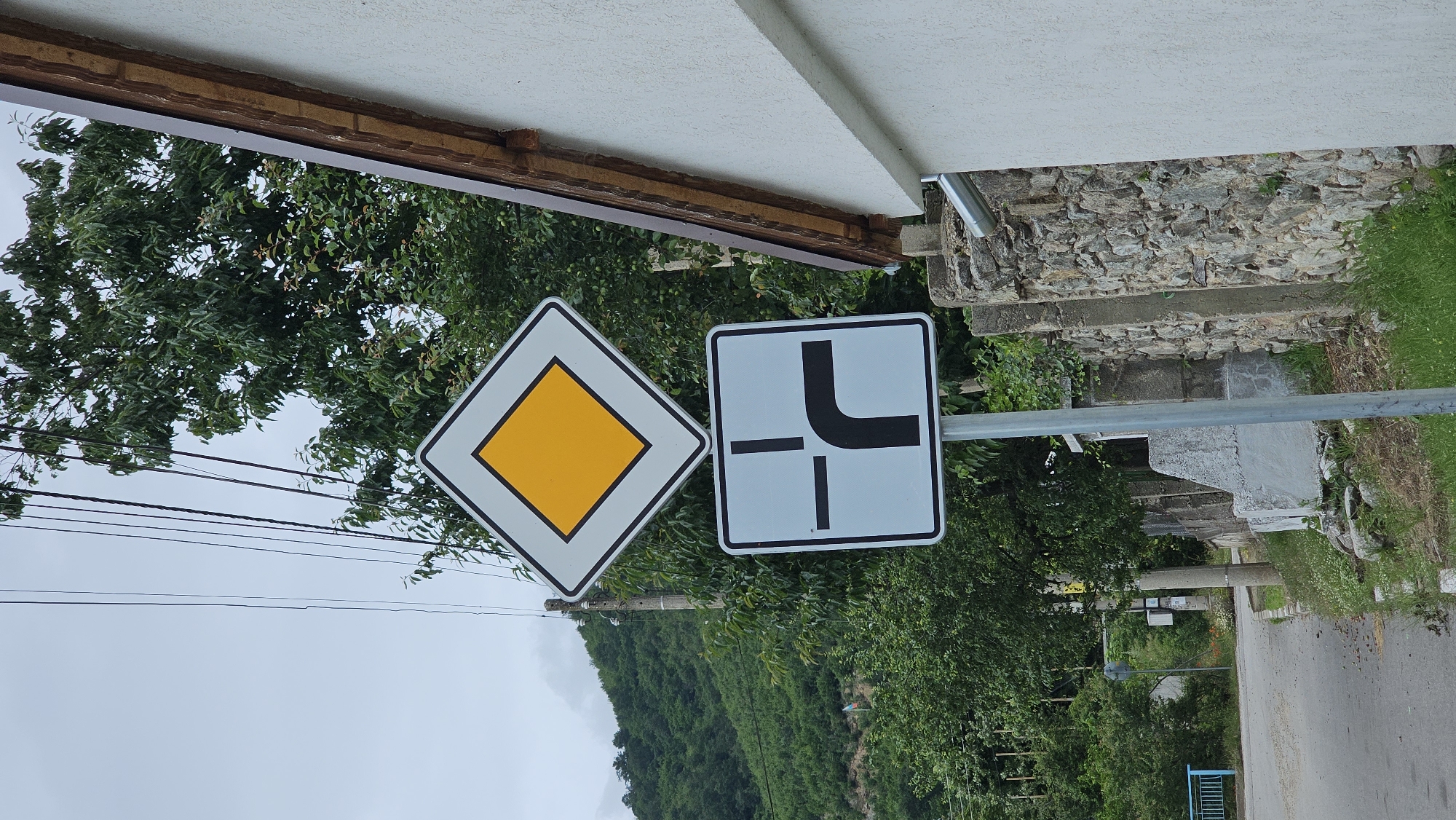 A diamond-shaped sign with a yellow interior and a white border, indicating a priority road, is mounted on a pole. Below it, a square sign with a black border and a light blue interior depicts a T-junction with a right turn from the side road. The signs are positioned next to a stone wall and a white building. The environment is a rural setting with trees and a cloudy sky.