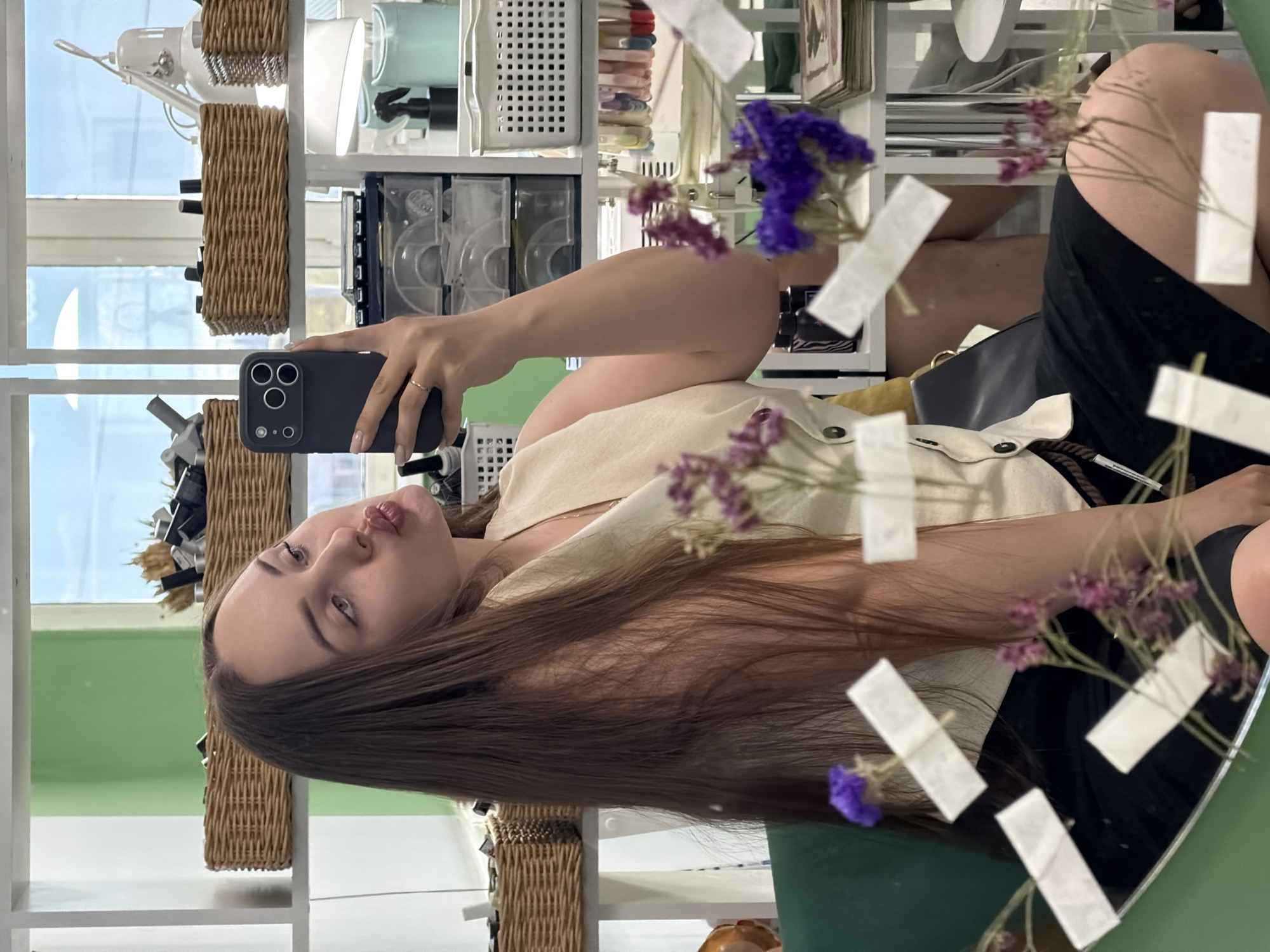 A young woman takes a selfie in a mirror. She has long brown hair and is wearing a light-colored top and dark shorts. The background shows shelves with various items, including wicker baskets and containers. There are also flowers and white pieces of paper taped to the mirror. The location is likely in Chaikovsky, Russia, given the prompt's instructions.