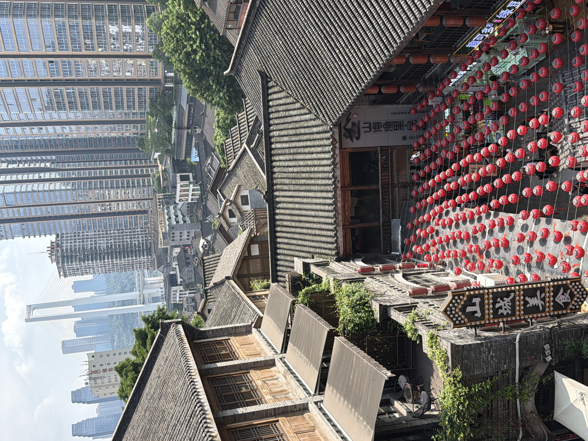 This is a high-angle, eye-level shot of an urban environment in Yuzhong District, China. The scene is a blend of traditional architecture and modern high-rise buildings. In the foreground, there are traditional Chinese-style buildings with grey tiled roofs. Some of these buildings have wooden balconies and awnings. A pathway is decorated with rows of red lanterns, suggesting a festive atmosphere. A vertical sign with Chinese characters is visible on one of the buildings. In the middle ground, more traditional rooftops are visible, interspersed with trees. In the background, a modern cityscape dominates, featuring numerous tall glass skyscrapers and a prominent cable-stayed bridge. The sky is partly cloudy. There are a few people visible walking along the lantern-lined pathway. The overall impression is of a city where old and new coexist.