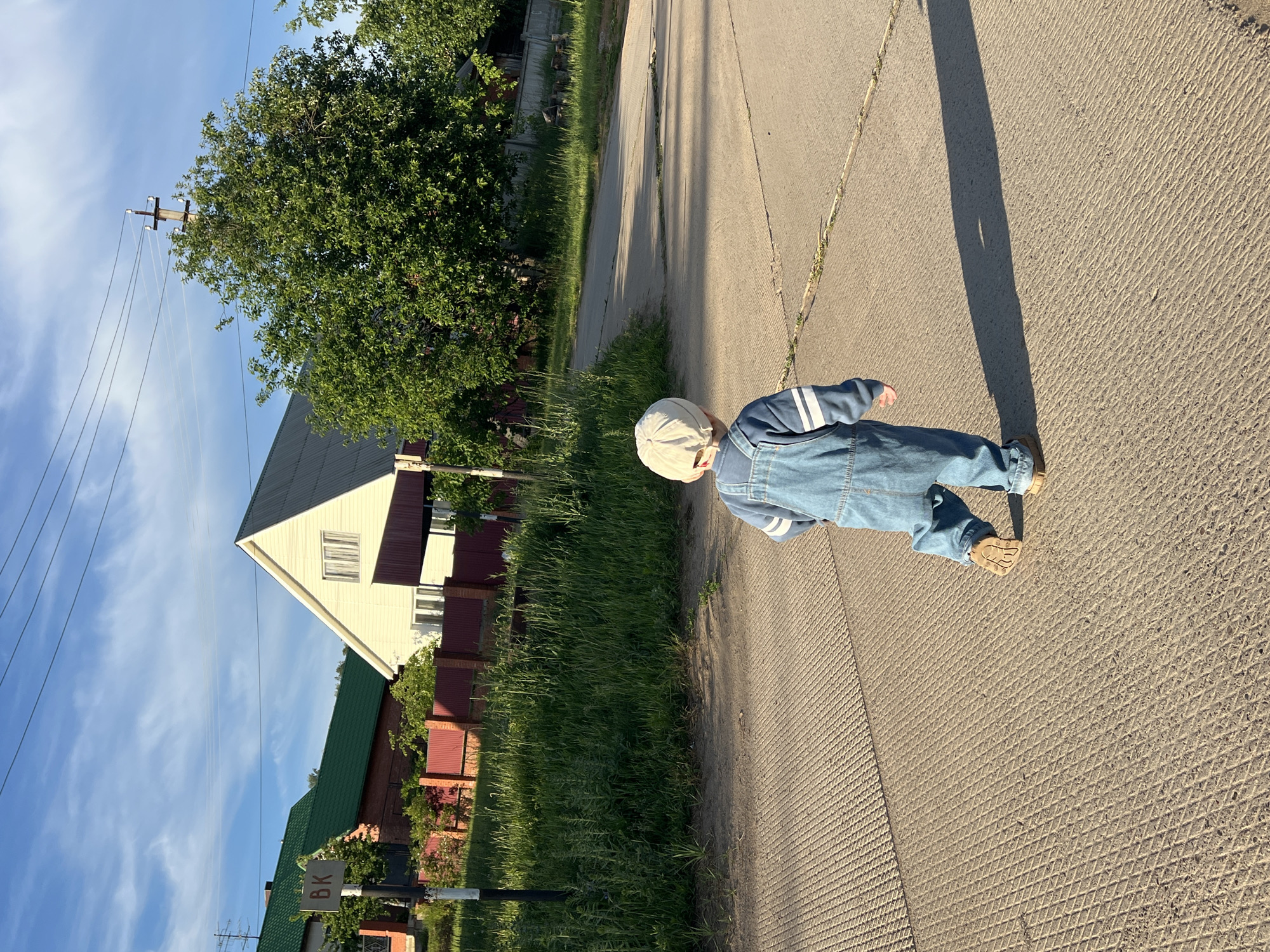 The image captures an outdoor scene featuring a young child from behind, walking on a textured concrete path. The child wears a light-colored cap, blue denim overalls, and a light blue long-sleeved top with white stripes on the sleeves. Their feet, clad in brown shoes, are visible near the bottom right.
The concrete path, characterized by a diagonal grid pattern, stretches from the foreground into the background. Long shadows are cast across the path, suggesting either early morning or late afternoon sunlight. To the left of the path, tall green grass grows alongside the road. Beyond the grass, several houses are visible, including one with light cream siding and a dark gray roof, and another with a green roof and brick walls, partially obscured by trees and red fences.
Lush green trees are scattered throughout the scene, particularly prominent on the left side and in the background. A utility pole with multiple power lines stands tall against the bright blue sky, which features scattered wispy white clouds. Near the bottom left corner, a signpost is partially visible with the white letters "BK" on a dark background, situated amongst the grass and a fence. The overall impression is a peaceful, sunny day in a suburban or rural setting.
