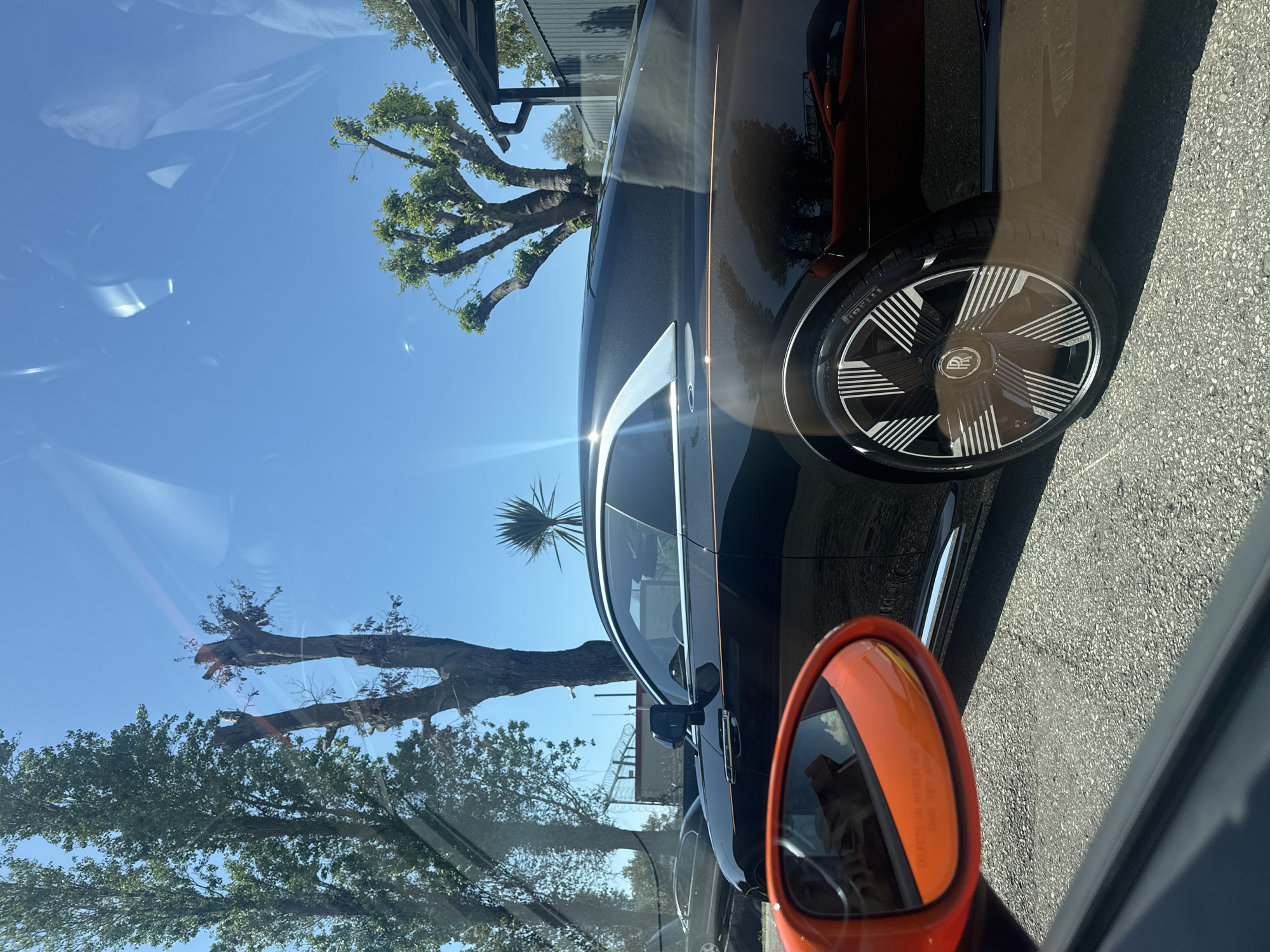 The image depicts a partial view of a dark-colored Rolls Royce, specifically its wheel and side, parked on an asphalt road on a bright, sunny day. The car's wheel has a distinctive design with radial lines emanating from the Rolls Royce logo. An orange-colored side mirror of another vehicle is visible in the foreground. The background includes trees and buildings. The location can not be definitively confirmed as Sochi, Russia, based on the visual information provided.