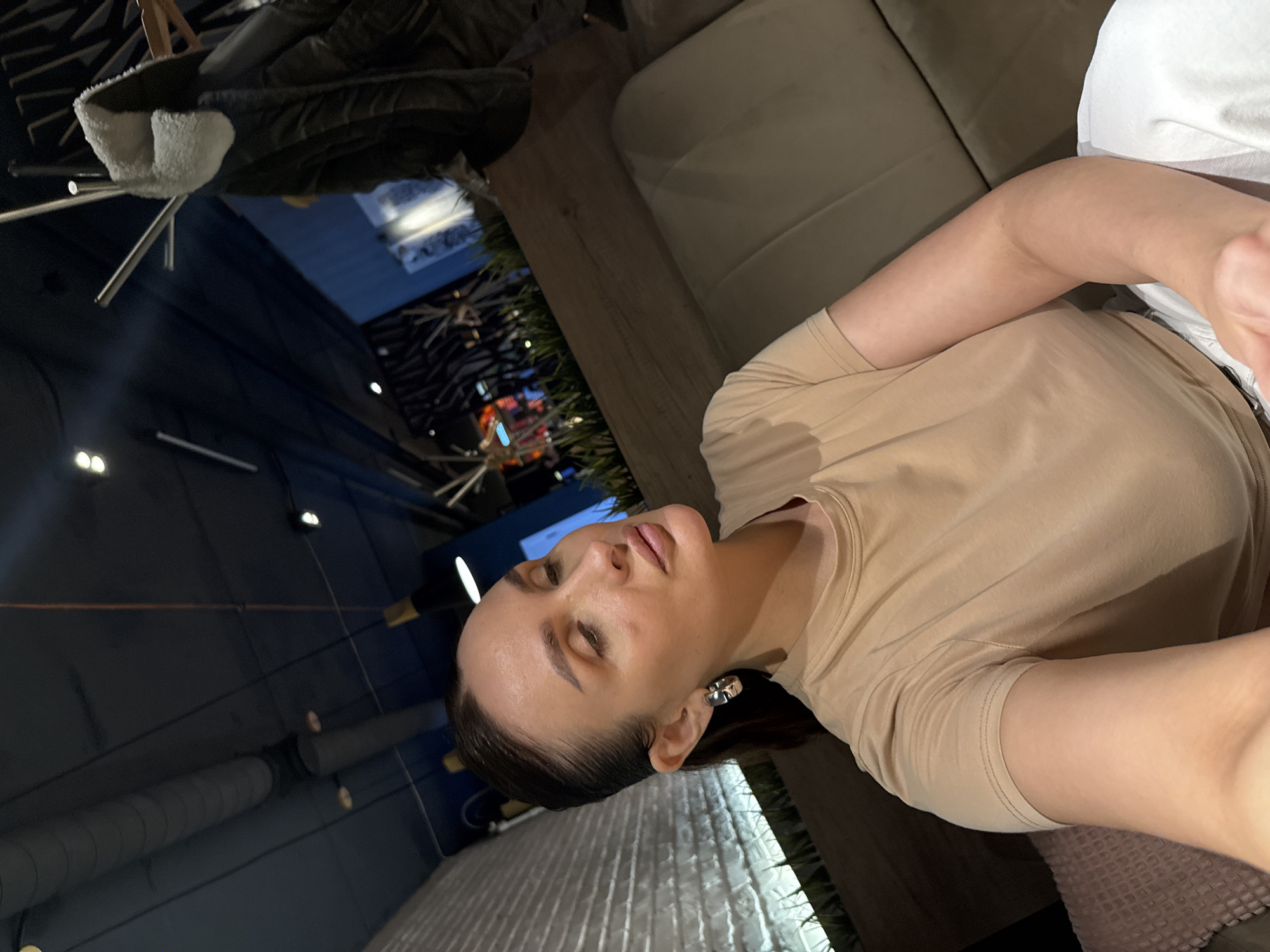 A woman is visible in a selfie-style image. She has dark hair pulled back and is wearing a light tan t-shirt. The background suggests an indoor setting, possibly a restaurant or lounge, with dimmed lighting and an urban cityscape seen through a window. The location is indicated as the city of Chaikovsky, Russia.