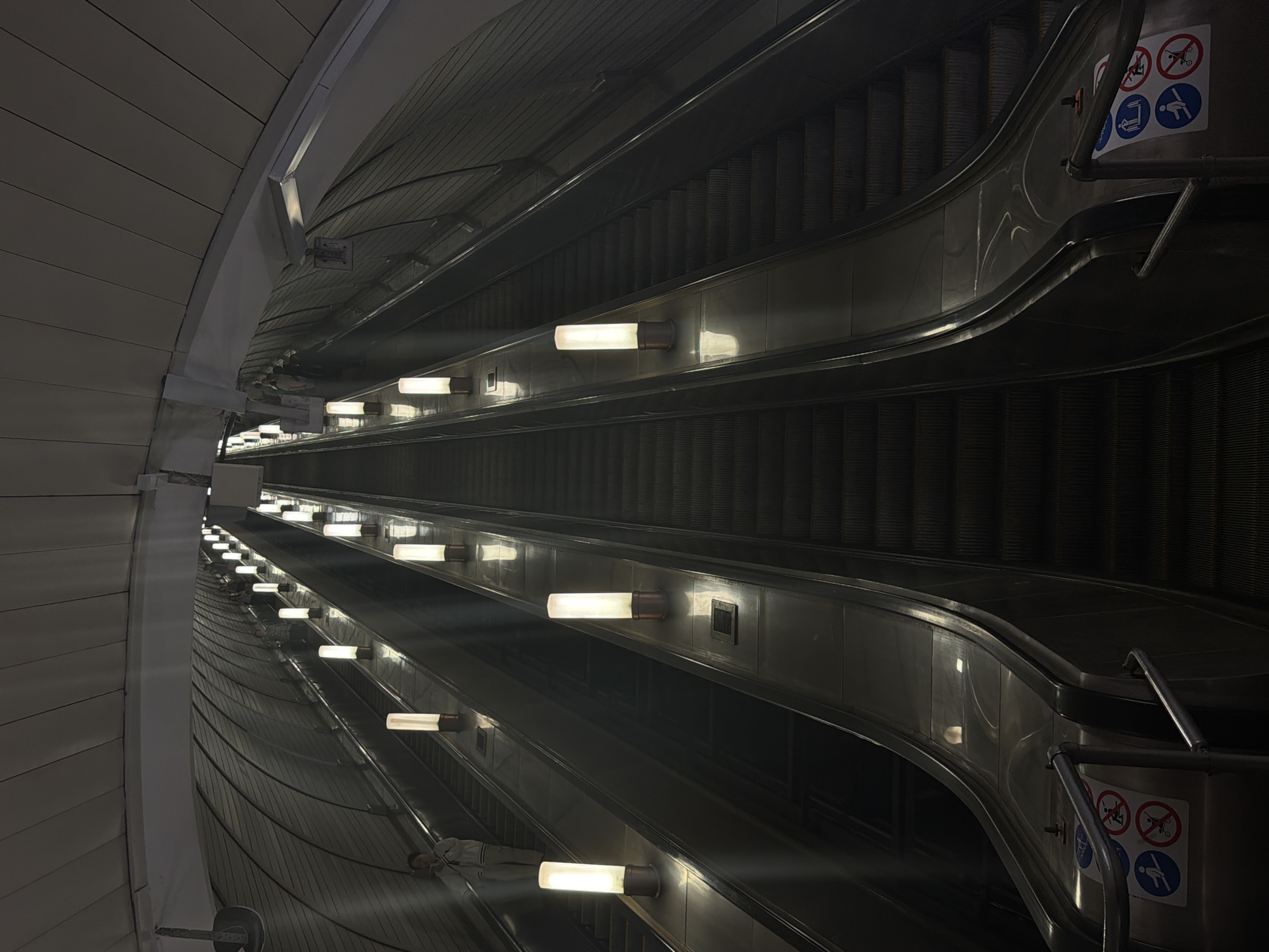 This image captures a low-angle, deep-perspective view of three parallel, very long escalators ascending into a dark, vast tunnel, characteristic of a deep-level Moscow Metro station in Moscow, Russia. The escalators are made of dark metal steps and shiny silver-grey side panels.
Bright, cylindrical light fixtures with a warm white glow are regularly spaced along the dividing panels between the escalators, casting vertical beams of light upwards and illuminating the surrounding structure. The tunnel's ceiling and upper walls are curved, ribbed, and dark grey, with lighter, arched structural elements visible towards the top.
On the leftmost escalator, a solitary figure, dressed in light-colored clothing, stands facing partially away from the camera, seemingly engaged with a small object near their face. Further up the rightmost escalator, several indistinct figures appear as dark silhouettes, also ascending. The middle escalator appears largely empty.
At the base of the escalators, silver metal handrails are visible, alongside warning signs affixed to the escalator's side panels. These circular signs include blue pictograms depicting a person holding a handrail, and red pictograms indicating "no running," "no luggage/strollers," and "no playing/sitting" on the escalator. The overall scene emphasizes the immense scale and depth of the underground transport system, with the lights providing a guiding pathway through the darkness.