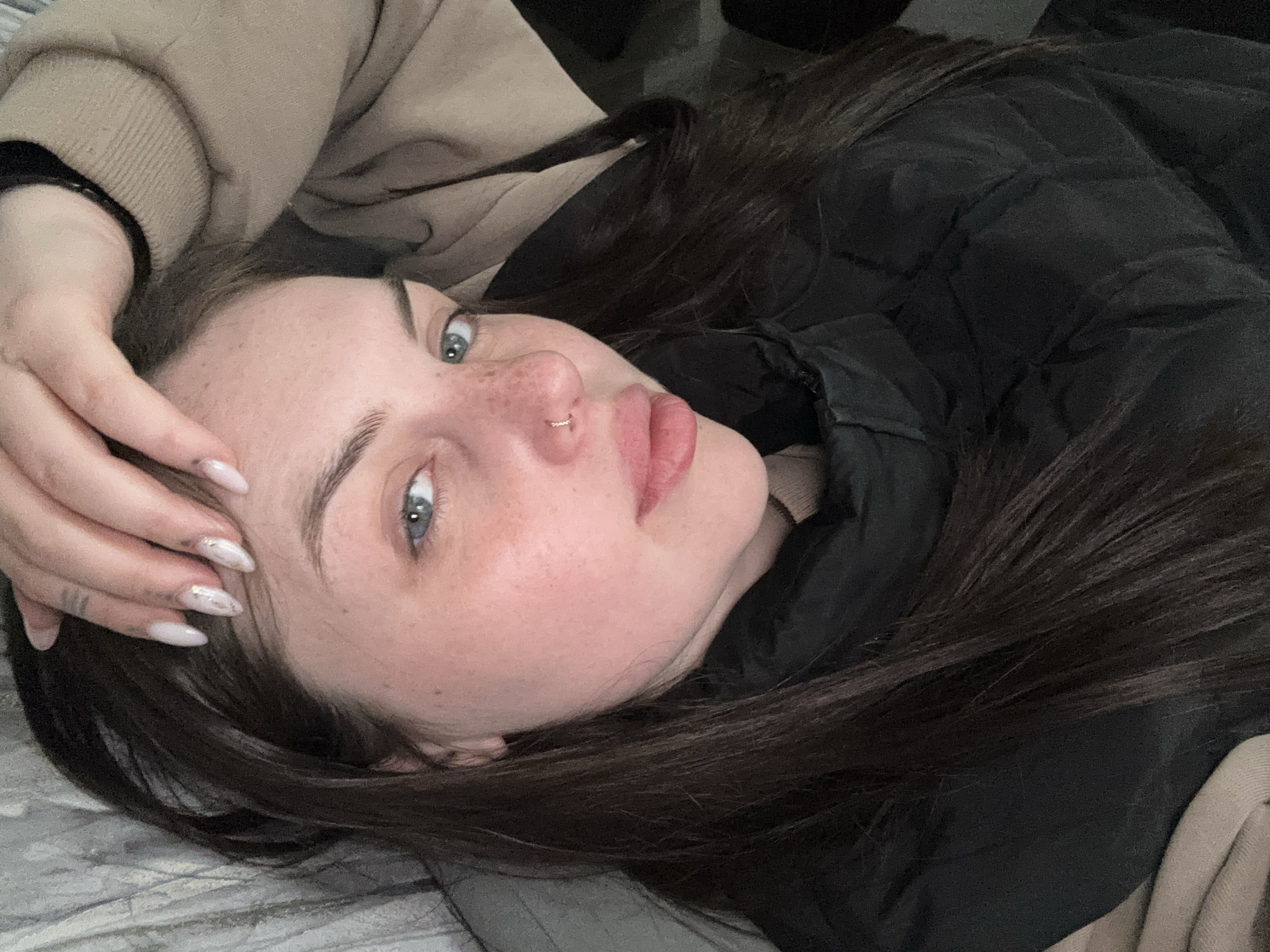 The image presents a close-up, angled portrait of a young individual, likely female, lying down indoors. Her head is tilted, and she is looking directly towards the camera.
**Subjects:** The central subject has long, dark brown hair that frames her face and spreads across the background. She possesses fair skin with scattered freckles across her nose and cheeks. Her eyes are a striking light blue-grey, framed by well-defined dark eyebrows. A small, golden hoop piercing is visible in her right nostril. Her lips are full. Her right hand is raised, with fingers gently resting near her forehead and hair. Her nails are long, almond-shaped, and appear to have a light-colored polish with subtle decorative elements, possibly glitter or a delicate pattern.
**Objects:** Key objects include the small golden nose ring and the manicured nails.
**Scene/Setting:** The setting appears to be an indoor space. The person is resting on a soft, textured surface, possibly a bed or couch, indicated by a grey, patterned fabric visible beneath her. A light beige or tan soft fabric, possibly part of her clothing or another blanket, is also visible in the background.
**Activity or event taking place:** The individual is captured in a relaxed pose, lying down and gazing directly at the viewer, suggesting a moment of quiet repose or an intentional photograph.
**Notable details:** She is dressed in a black quilted or puffer jacket, visible on her left side, layered over a lighter, possibly beige or tan, garment. Her expression is neutral and calm. The lighting suggests an indoor environment. There are no specific cues within the image to indicate the time of day or external weather. No location-specific elements are visible in the photograph that directly relate to Chornomorskoe, Ukraine.
**Visible text:** There is no visible text in the image.