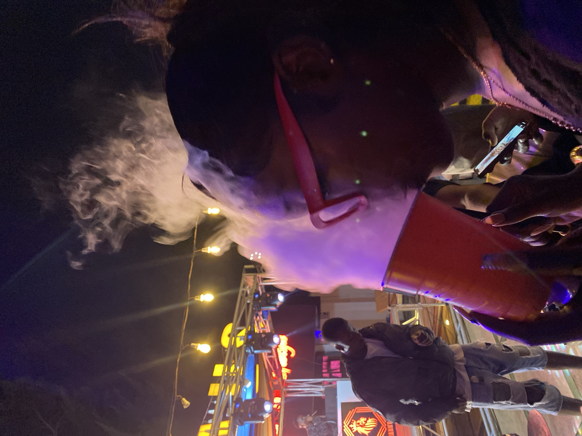 The image depicts a nighttime outdoor scene, possibly a music venue or festival, with a stage and lighting setup in the background. In the foreground, a person in profile is holding a red cup, from which a plume of smoke or vapor is emanating. The person is wearing red-rimmed glasses and a dark top, with a necklace visible. A smartphone is also held in their other hand. To the left, another person is standing near a DJ setup, wearing a dark jacket and ripped denim shorts. The overall atmosphere appears to be lively, with string lights illuminating the area.