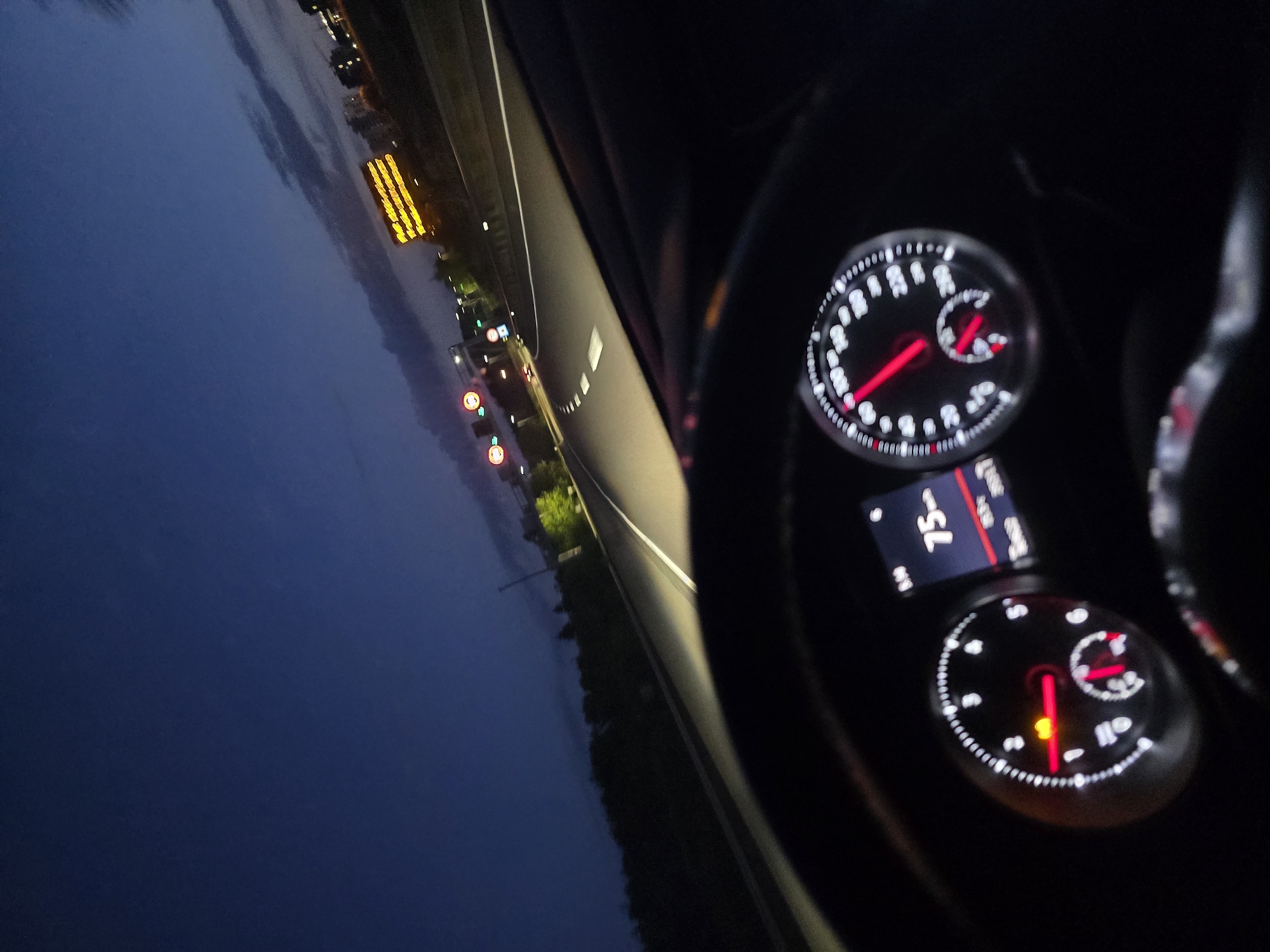 The image is a first-person perspective shot from inside a car driving on a highway at dusk. The dashboard is visible in the foreground, showing a speedometer indicating a speed of 75 km/h. The car's headlights illuminate the road ahead. In the background, the sky is a deep blue, transitioning to lighter hues near the horizon with some clouds. There are streetlights and signs visible along the side of the highway. One prominent digital sign displays text in yellow. The location is identified as Russange, France. The scene depicts a common driving scenario on a highway.