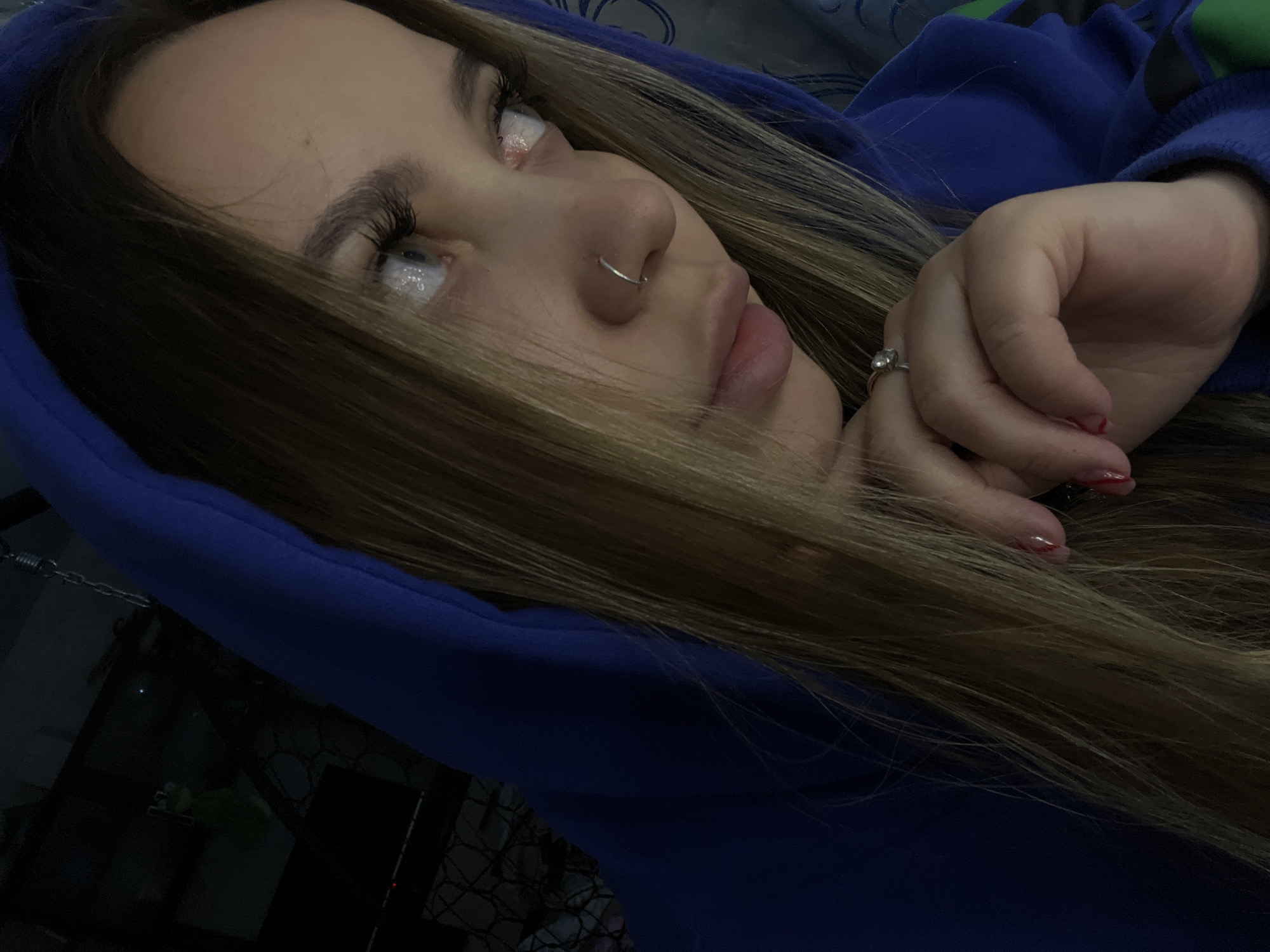 A close-up, slightly angled photograph shows a young woman wearing a blue hooded sweatshirt. She has long, light brown hair and is wearing a nose ring. Her left hand is near her face, displaying a ring on one of her fingers and nail polish on her fingernails. The background is dark and indistinct, but a television and some other objects can be seen in the lower left corner. The location is unspecified and the city of Novopokrovskaya, Russia, is irrelevant to the visual content of the image.