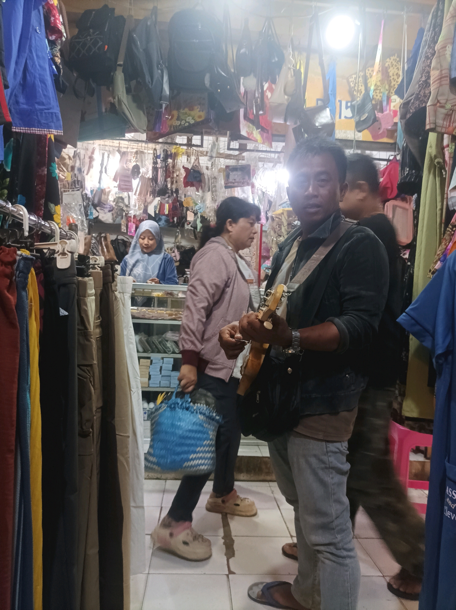 The image depicts an indoor scene, likely a market stall in Sumuragung, Indonesia, filled with merchandise. In the foreground, a man wearing a dark jacket over a grey t-shirt and blue jeans is playing a guitar. He has a guitar strap slung across his chest and a black bag. To his left, a woman wearing a blue headscarf and a blue top is visible behind a counter with shelves stocked with items. To the right of the man, another person, whose face is obscured, is walking past. The background is filled with various items hanging and displayed, including bags and clothing. The floor is tiled. The lighting suggests an artificial light source overhead.