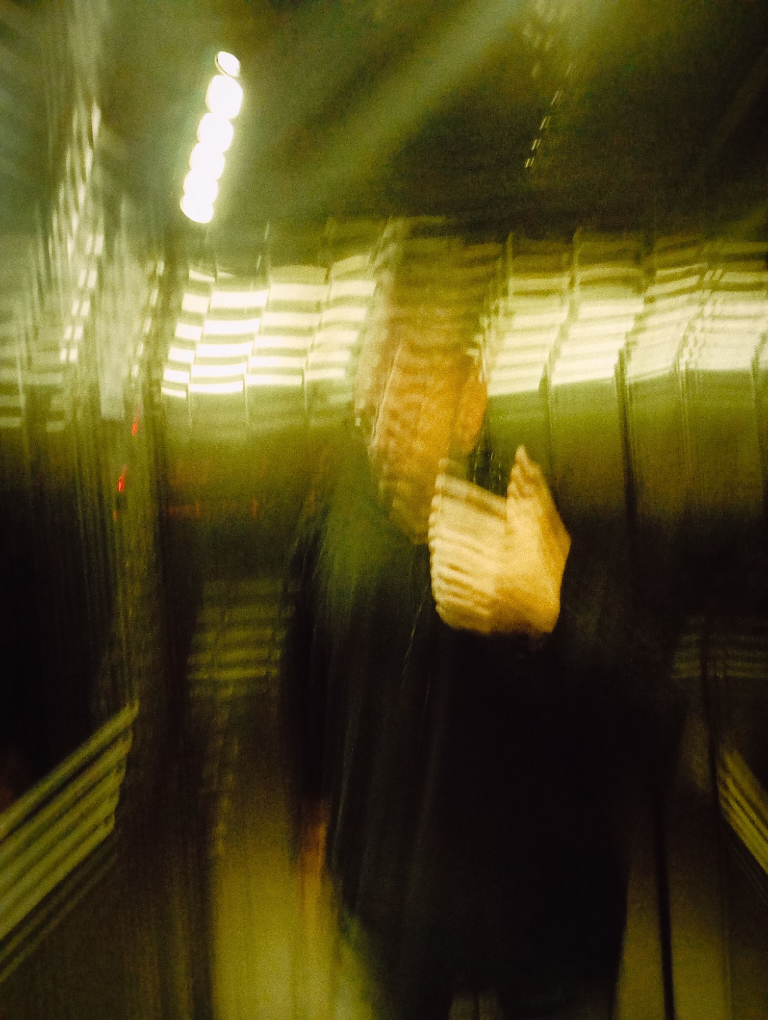 The image is a blurry, abstract depiction of an interior, possibly an elevator, with a person inside. The lighting is artificial and dim, with a row of lights visible at the top. The person appears to be wearing a dark-colored shirt or jacket and is gesturing with a hand. Due to the blur, detailed identification of objects and environment is impossible. There is no discernible information in the image to identify a location in Buenos Aires, Argentina.