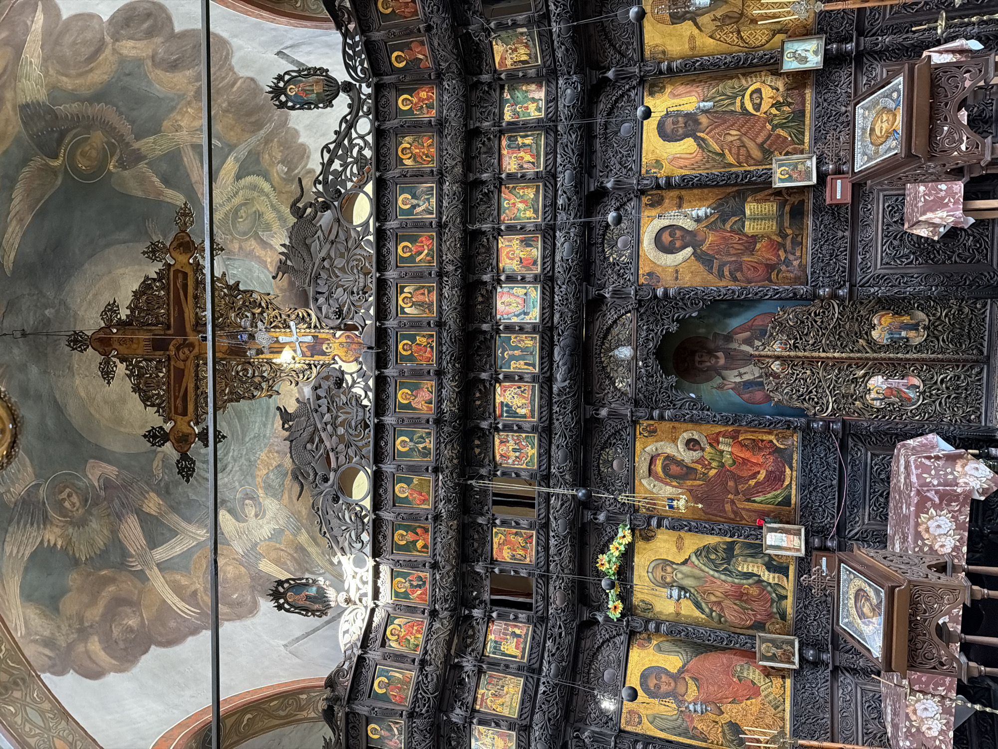 This image captures an ornate ceiling within a religious building, likely a church or monastery, in Sevlievo, Bulgaria. The ceiling is intricately decorated with both painted frescoes and numerous religious icons set within dark, carved wooden panels.
The frescoes on the left depict winged figures, likely angels, in muted tones of brown and grey, set against a cloudy sky. Below the frescoes, a large wooden cross with ornate carvings is visible.
The majority of the ceiling is covered by rows and columns of small, rectangular icons, predominantly featuring religious figures such as saints and what appear to be depictions of the Virgin Mary and Jesus Christ. These icons are in a traditional Byzantine or Eastern Orthodox style, characterized by their gold backgrounds and stylized figures. Larger icons are also present, with detailed paintings of religious scenes and figures. Small, decorative wooden elements and hanging lights are interspersed throughout the ceiling.
The overall impression is one of solemnity and religious devotion. The detailed artwork and the sheer number of icons suggest a place of significant spiritual importance. The lighting appears to be a mix of natural light filtering in and possibly artificial lighting, creating a somewhat dramatic effect on the rich textures and colors of the artwork. No specific time of day or weather conditions are indicated. There is no visible text within the image itself, beyond what might be present on the small icons, which is not legible at this resolution. The setting is clearly an interior religious space, with its characteristic architectural and artistic elements.