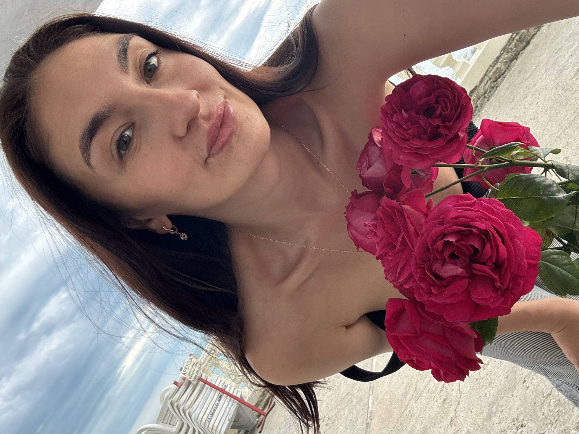 A young woman is posing for a selfie on the beach in Sochi, Russia. The woman has long brown hair and is wearing a small gold necklace and earring. She is holding a bouquet of large, red roses. In the background, there is a view of the beach, sky, and white stacked beach chairs with Russian text on a sign.