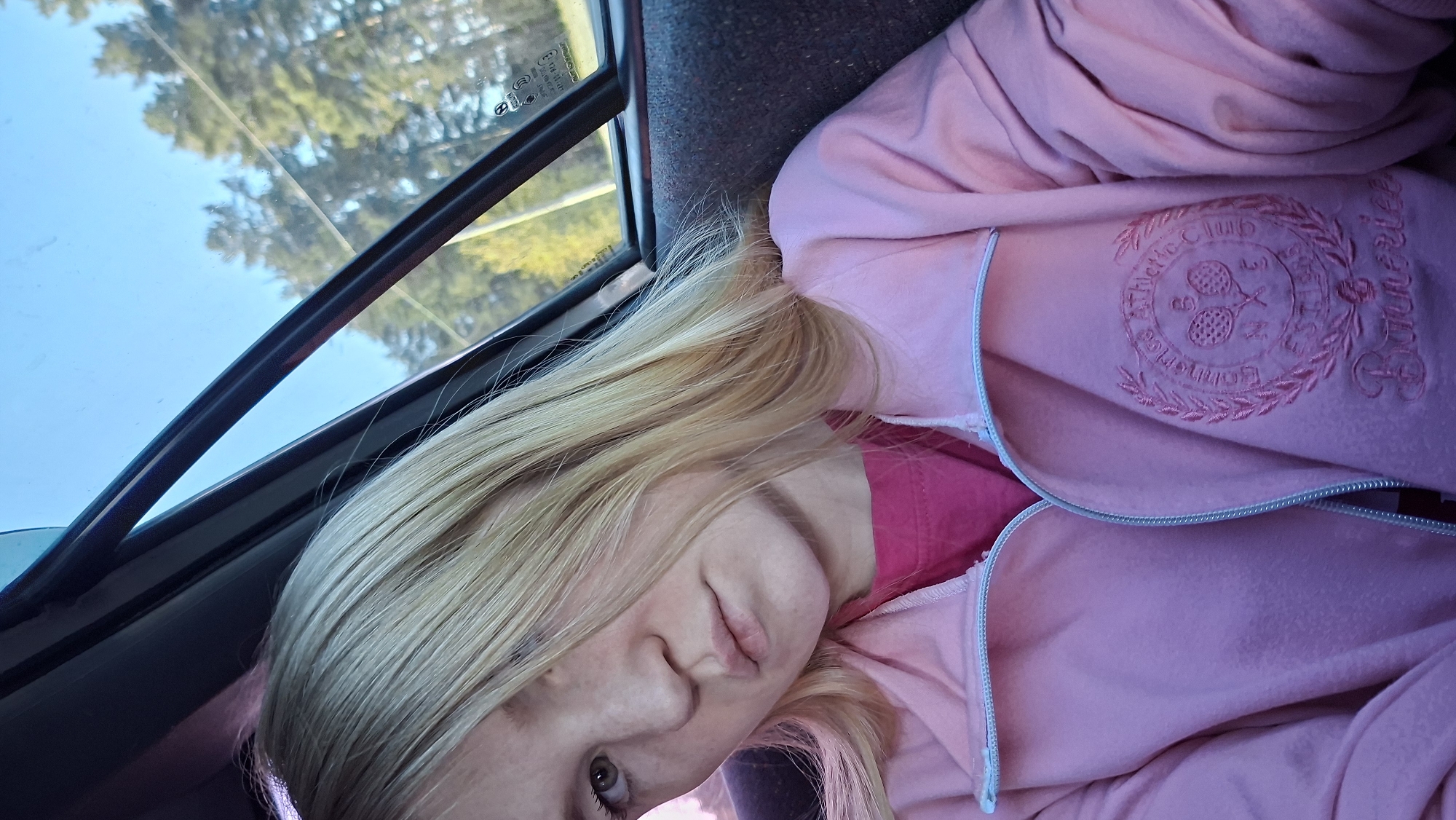 A fair-skinned person with blonde hair is depicted in a close-up, rotated view. They are wearing a pink zippered jacket over what appears to be a pink shirt. The jacket has an embroidered logo on the upper right side with the words "Athletic Club" and "Bonarice." The person is positioned near a window with a view of trees and the sky outside. The location details of Chaikovsky, Russia, are not directly discernible from the image.