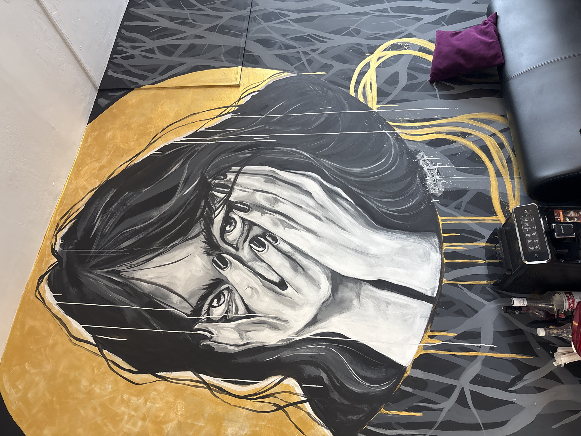 The image shows a mural depicting a woman covering her face with her hands. The mural is predominantly black and white, with accents of gold and gray. The woman has dark hair, and her eyes are visible between her fingers. The background of the mural is a mix of abstract shapes and lines in black, gray, and gold. A purple pillow and a black leather chair with a remote control on it sit near the mural. An empty bottle of Barmatoff Vermouth and napkins are also visible.
Based on the artist's signature, the mural is by Carl Grodanva. The location is likely within a business in Krasnoyarsk, Russia.