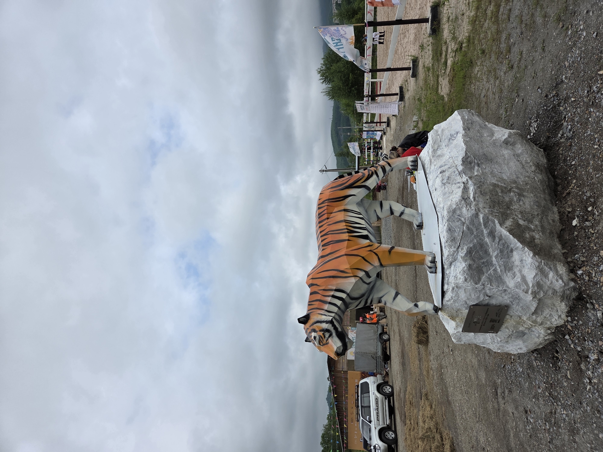 An outdoor scene in Теплоозёрск, Russia features a large, geometric tiger sculpture prominently displayed on a light-colored, rough-textured boulder. The tiger is orange and black with angular stripes, and appears to be walking on a flat, metallic surface. In the background, a silver SUV with "PRADO" branding is parked to the left. Behind it, a covered trailer and a low wooden building with flags and banners are visible. Several people are present, including one wearing an orange vest near the trailer and others in the distance. The ground is covered in gravel and dry grass, with green grass growing in patches. The sky is overcast with clouds, and hills with trees are visible in the distance.