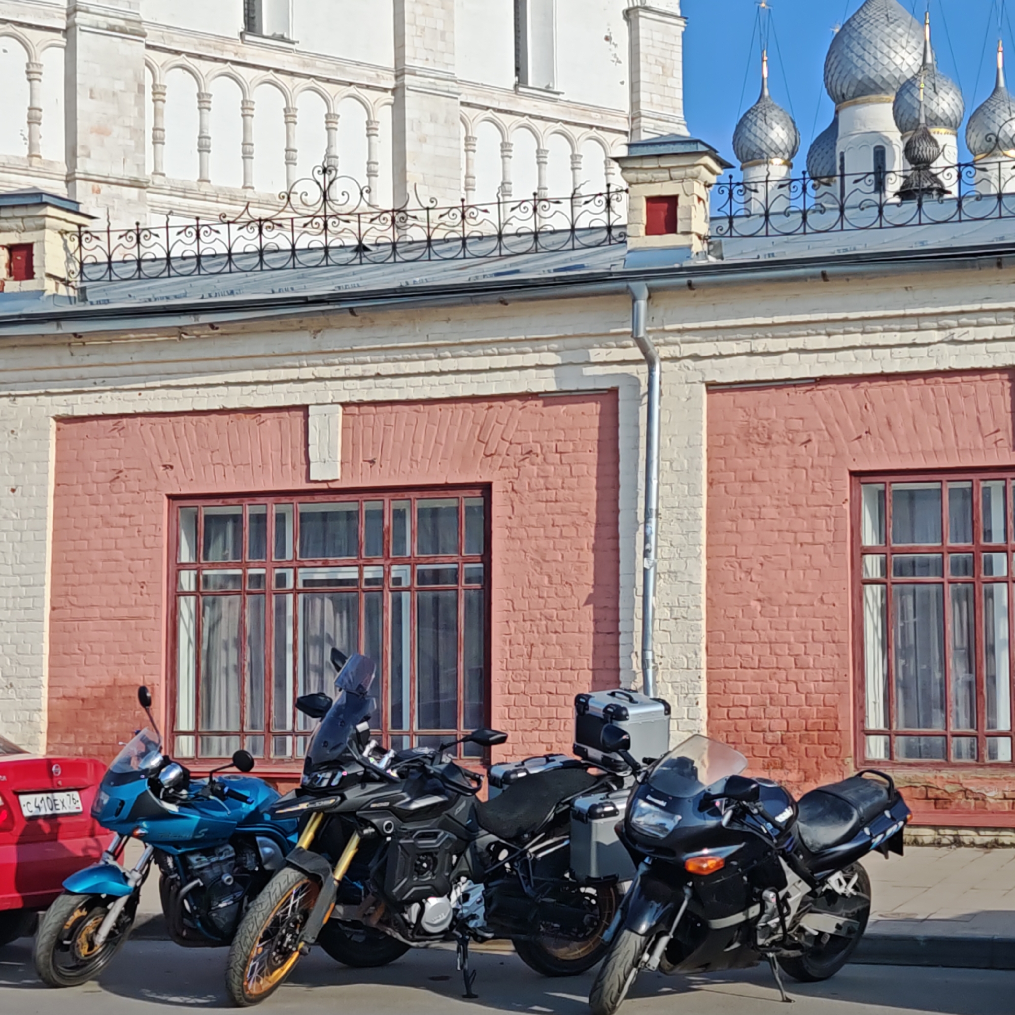 The image depicts a street scene in Rostov, Russia, featuring four parked motorcycles in front of a two-story building with pink brickwork and white trim. The building's architecture includes decorative ironwork along the roofline and silver onion domes visible in the background. A small portion of a red car is visible on the left edge of the frame. The motorcycles are various colors and models, including a blue Suzuki, a black adventure bike, and two black Kawasaki models. The presence of the motorcycles suggests a gathering or parking area for riders. The overall scene appears to be a quiet street with a combination of historical architecture and modern vehicles.