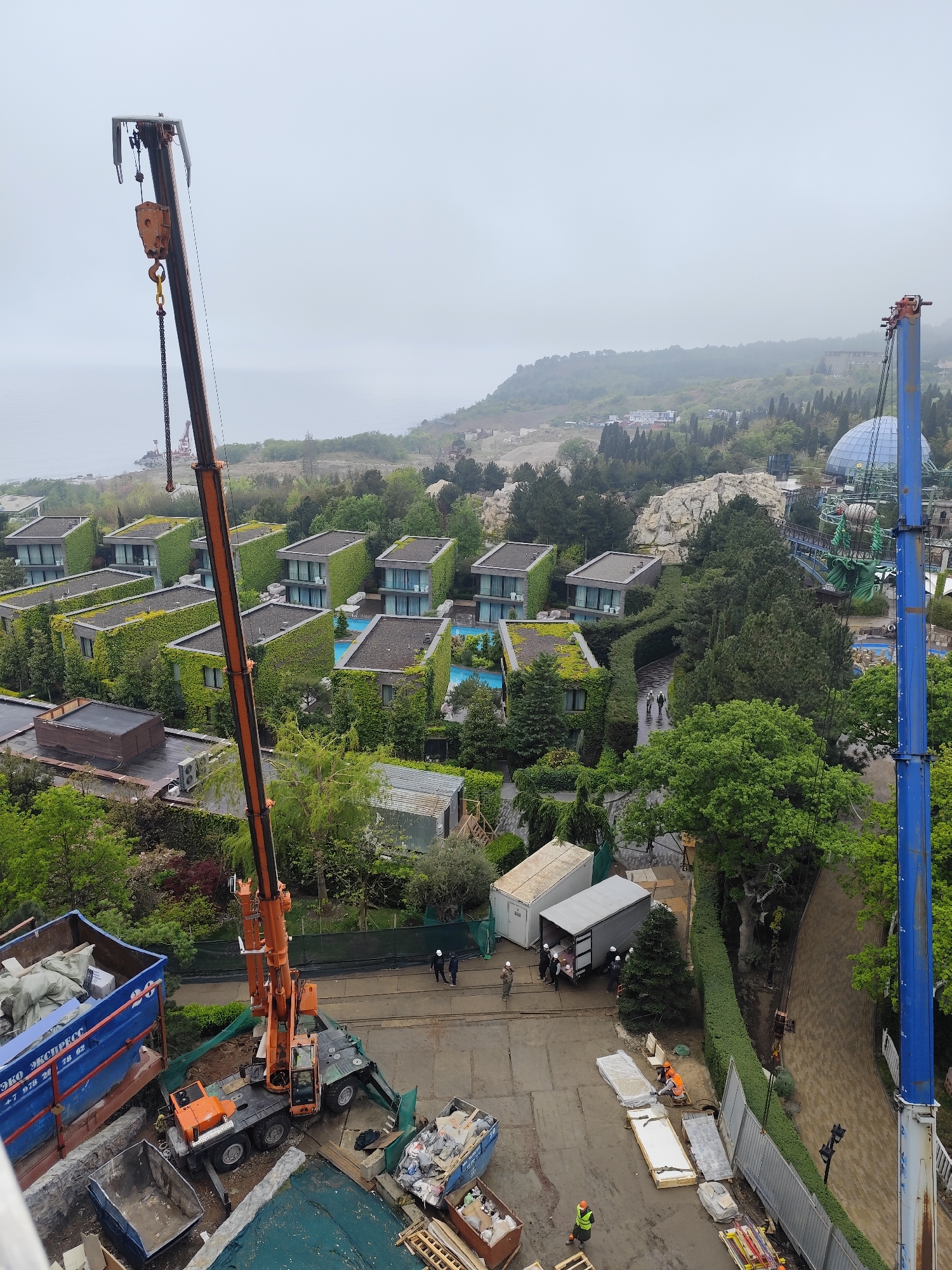 Captured from an elevated perspective, the image portrays a construction site in Симеизский поселковый совет, Ukraine. The landscape encompasses structures with verdant, plant-covered roofs adjacent to recreational areas featuring swimming pools and thematic park elements such as a blue geodesic dome and a green octopus-like ride.
In the foreground, an active construction zone is evident. An orange crane dominates the scene, positioned next to a blue refuse container filled with construction debris. Further towards the bottom of the frame, a smaller dumpster overflowing with waste and discarded materials is also visible. A construction crew, some wearing hard hats and safety vests, are engaged in various tasks. Some are clustered around a white truck and a smaller trailer, possibly unloading or organizing materials. Others are kneeling near a stack of rectangular white panels, seemingly preparing them for installation. A green tarp and scattered wooden planks add to the construction clutter.
Beyond the immediate construction area, a meticulously maintained green hedge lines a pathway, suggesting a resort or high-end residential development. Further in the background, the scene blends into a wooded landscape transitioning to a hilly terrain under a clouded sky. The atmosphere appears overcast, typical for the coastal region.