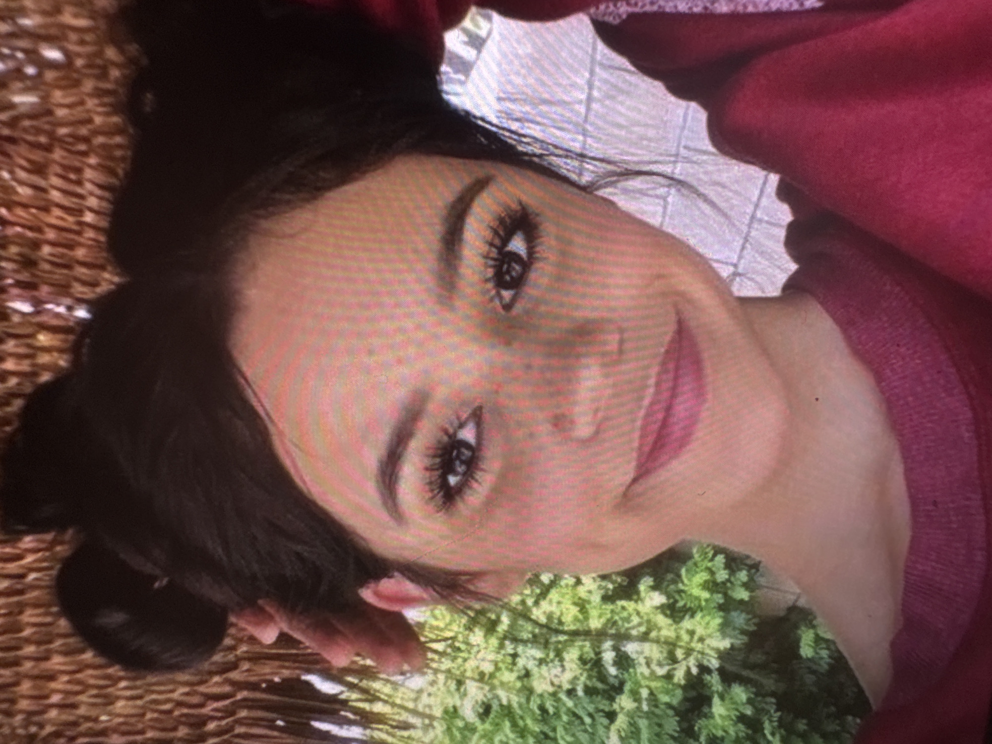 The main image features a young woman with dark hair and fair skin, smiling gently at the camera. Her head is angled towards the top right of the frame, giving the impression that she is reclining or the photograph was taken from an overhead perspective. She has prominent, well-defined eyebrows, long dark eyelashes, and is wearing pink lipstick. Her dark hair appears to be pulled back or styled, with some strands framing her face. She is wearing a red or maroon long-sleeved garment, possibly a sweatshirt or sweater.
The setting appears to be outdoors during the daytime, suggested by the natural lighting on her face and the vibrant green foliage visible in the background, particularly towards the bottom and right side of the image. On the left side, a textured, woven brown material, resembling wicker or rattan furniture, is visible. The woman's expression conveys a sense of calm and happiness. There is no visible text in the image.