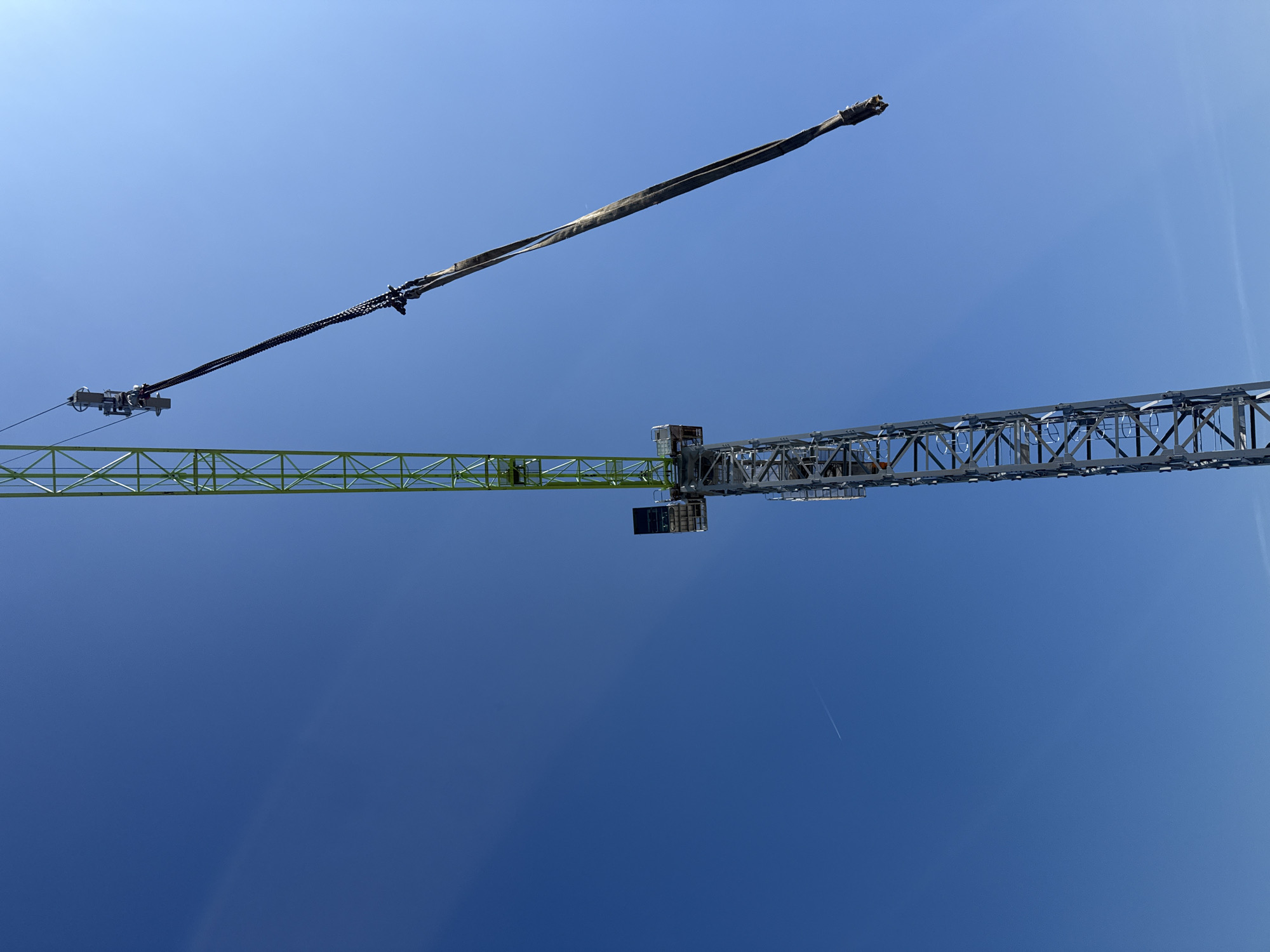This image captures a worm's-eye view of a large tower crane set against a clear, deep blue sky in Varna, Bulgaria. The crane's structure dominates the upper portion of the frame, showcasing its intricate metalwork.
The primary subjects are the various components of the crane. To the left, a vibrant light green section of the crane's jib extends horizontally, characterized by its lattice-like construction. Running along this jib, and then continuing diagonally upwards towards the top center, is the main hoist rope, which appears thick and wrapped in a protective covering in some sections, with a complex trolley mechanism visible towards the left end of the jib.
To the right, a darker grey or silver section of the crane's structure, likely part of the counter-jib or mast, also features a robust lattice design. A small cabin or platform is visible at the junction of the green and grey sections. The overall scene suggests an active construction site, though no specific lifting activity is captured, and no people are visible.
Notable details include the bright daylight, indicated by the vibrant blue of the uninterrupted sky, suggesting sunny and clear weather conditions. A faint, thin white airplane contrail is visible in the lower right quadrant of the sky, adding a subtle element of distant air traffic. The stark contrast between the industrial crane and the expansive natural sky is a prominent feature of the composition. No discernible text is visible in the image.