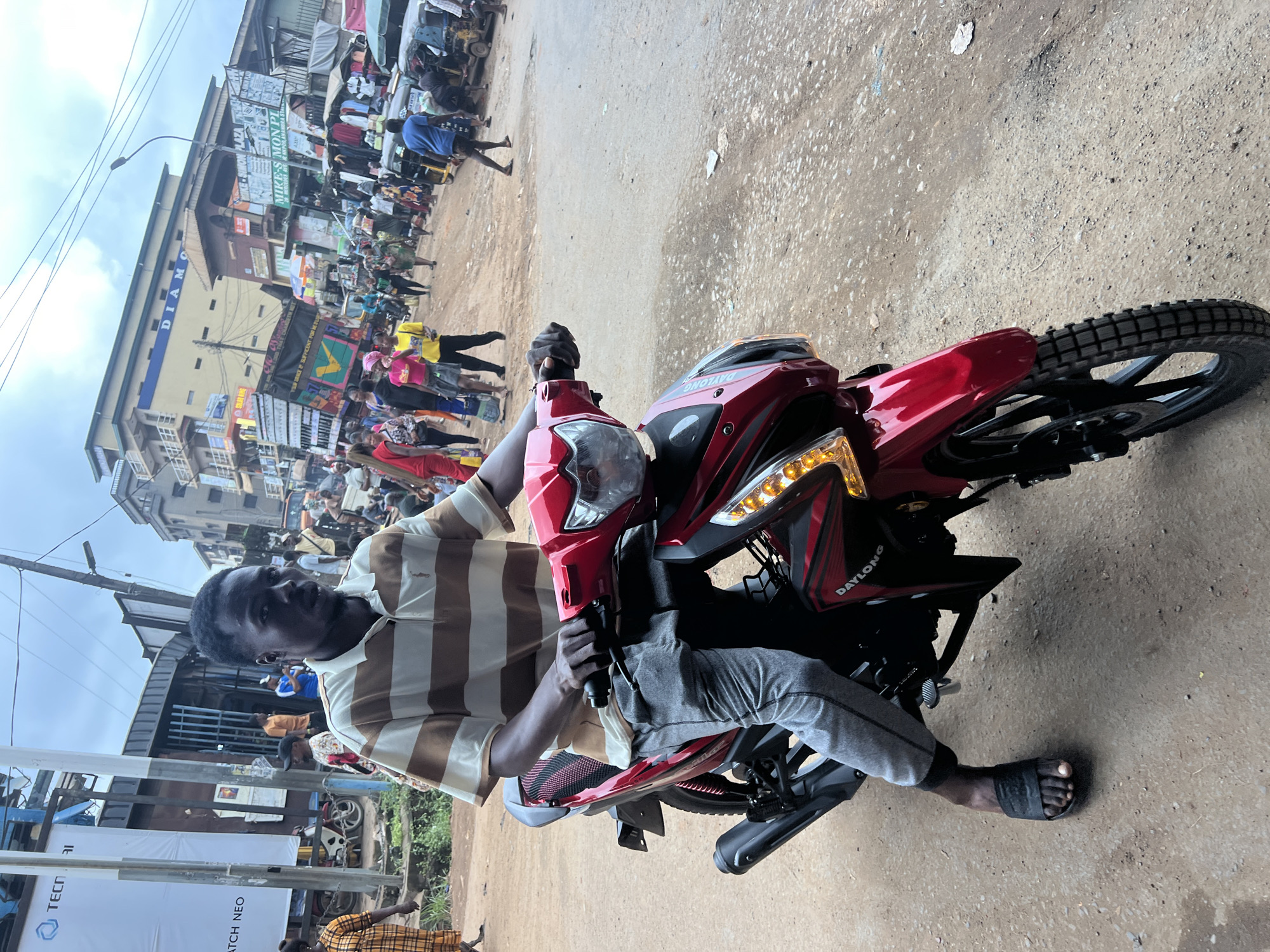 A young Black man wearing a brown and white striped polo shirt and grey sweatpants is seated on a red and black motorcycle. The motorcycle has a black exhaust pipe and black rims, and the brand "DAYLONG" is visible on the side. The man is looking forward, and his hands are on the handlebars. He is wearing black slide sandals on his feet.
The scene appears to be a busy street in Nkpor, Nigeria. The road is unpaved and dusty, with scattered debris. In the background, there are several buildings, including a yellow building with "DIAMOND" written on it, and other structures that appear to be shops or market stalls. There are many people walking on the street, some carrying goods, and several other vehicles, including a trishaw and a truck. The sky is overcast. The overall impression is that of a vibrant, bustling market area.