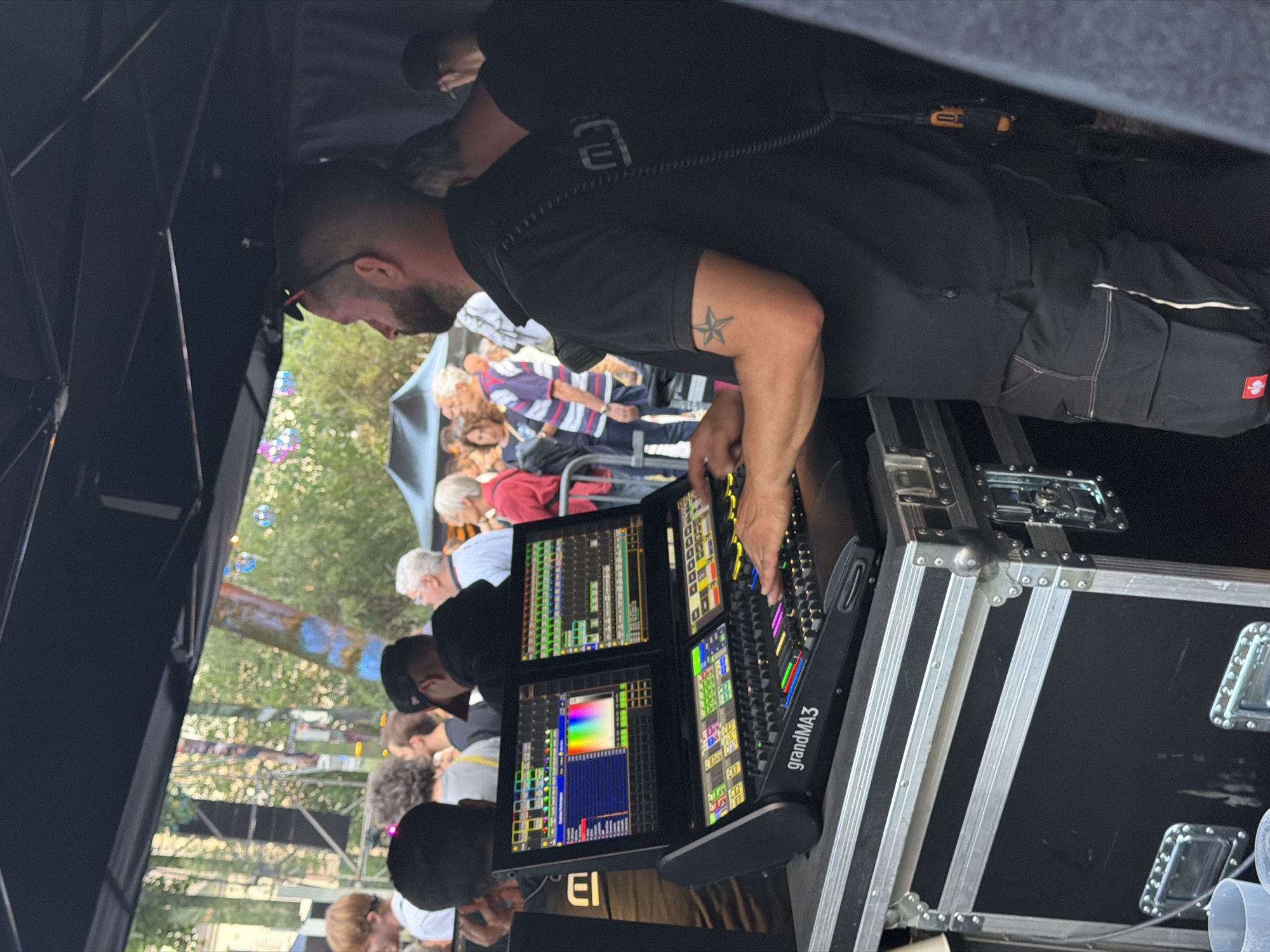A man, identified as a sound technician, operates a GrandMA3 lighting and show control console. He is standing under a black awning, presumably at an outdoor event. The console has two large screens displaying complex graphics and numerous faders and buttons, many of which are illuminated. The man's hands are on the console, suggesting he is actively controlling the show. He is wearing a black polo shirt and black cargo pants. A star tattoo is visible on his left forearm. In the background, a crowd of people are visible, some of whom are blurred due to motion. The environment appears to be a public space with trees and buildings visible in the distance. The event is likely taking place in Luxembourg City, Luxembourg.
