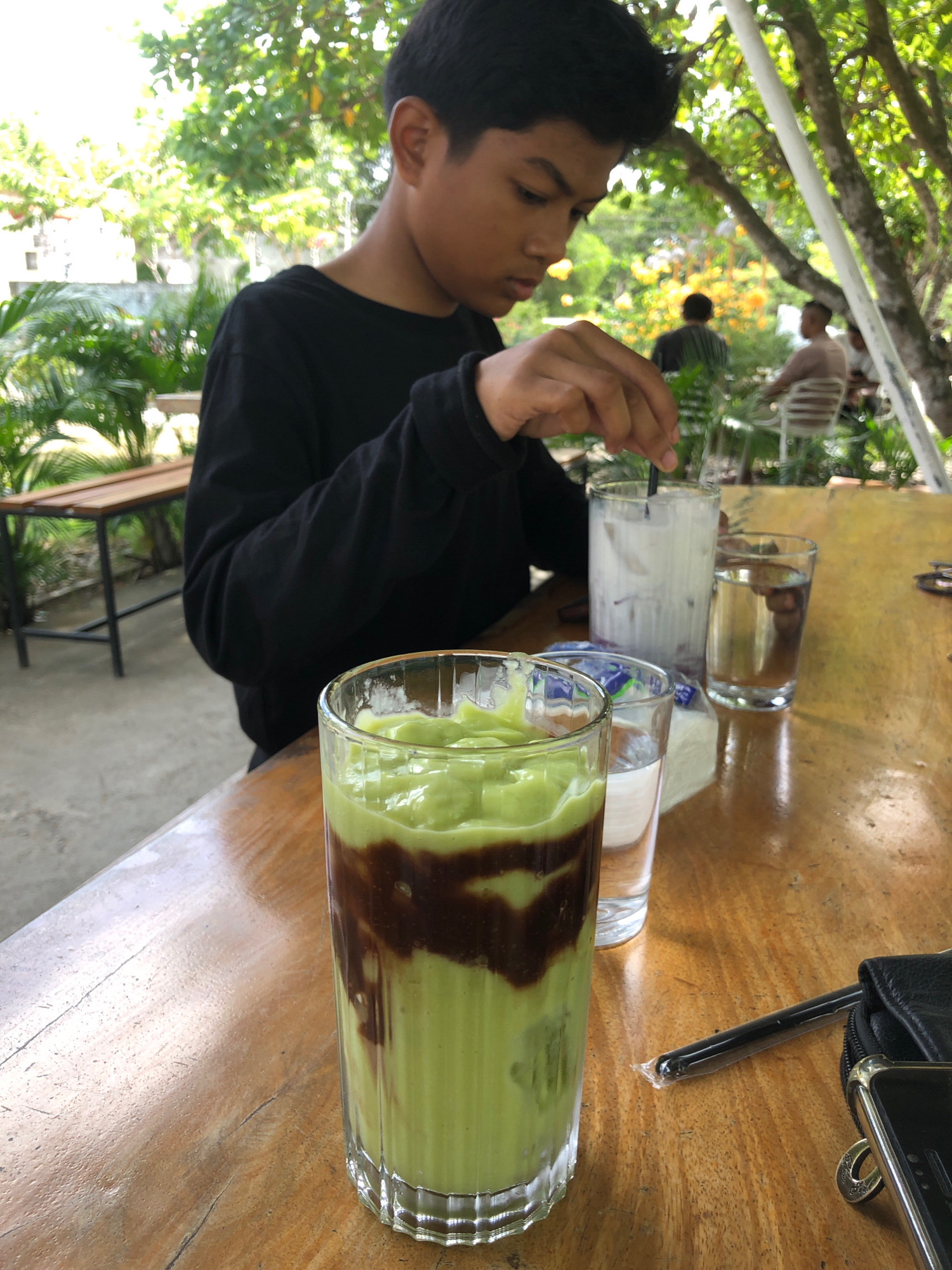 A young male, likely of Indonesian descent, is seated at a wooden table outdoors, with a glass of avocado smoothie topped with chocolate syrup in the foreground. He is wearing a black long-sleeved shirt and appears to be stirring a second drink, a tall glass of white liquid, with a straw. Behind him, two other individuals are visible in the background, seated at tables under a canopy of trees and foliage, suggesting a cafe or restaurant setting. The environment is lush and green, with palm trees and flowering plants. A black wallet and a smartphone are also on the table. The scene appears to be a casual outdoor dining experience in Tambonbaroh, Indonesia.