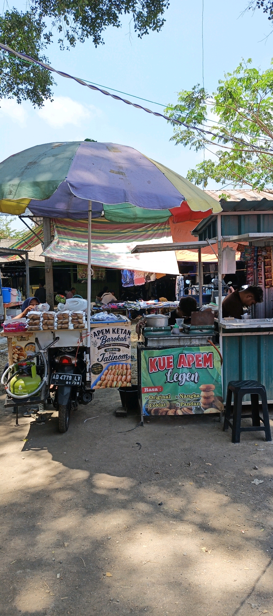 A street food stall is set up under a large, multi-colored umbrella in Jatinom, Indonesia. The stall features a variety of food items, including apem ketan and kue apem legen, with different flavors like original, nangka, coklat, and pandan advertised. A motorcycle is parked to the side of the stall, equipped with a gas cylinder. Several people are visible, some attending to the food and others seated. The ground is unpaved and dusty, with shadows cast from trees and overhead structures. The overall scene depicts a bustling outdoor market or roadside food vendor.