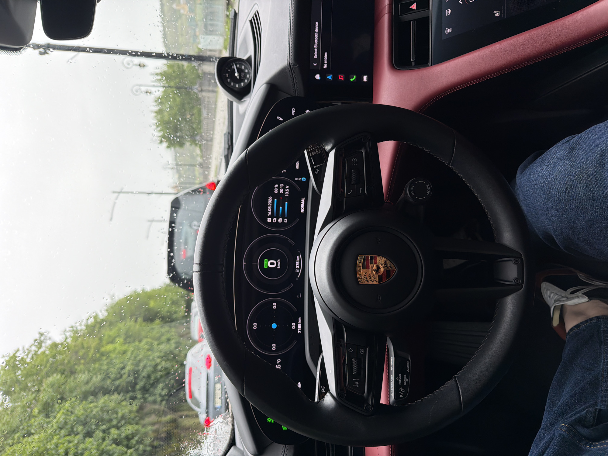 The image shows the interior of a Porsche, specifically the steering wheel and dashboard, from the driver's perspective. The dashboard indicates the car is not moving (0 km/h) and has a mileage of 7185 km. The date on the display is 16.05.2026. The steering wheel features the Porsche logo.
Outside the windshield, it is raining, and other vehicles are visible, suggesting the car is in traffic. Lush green trees are also visible. There is no information to identify the location as Tbilisi, Georgia. The image shows a driver wearing jeans and a sneaker.