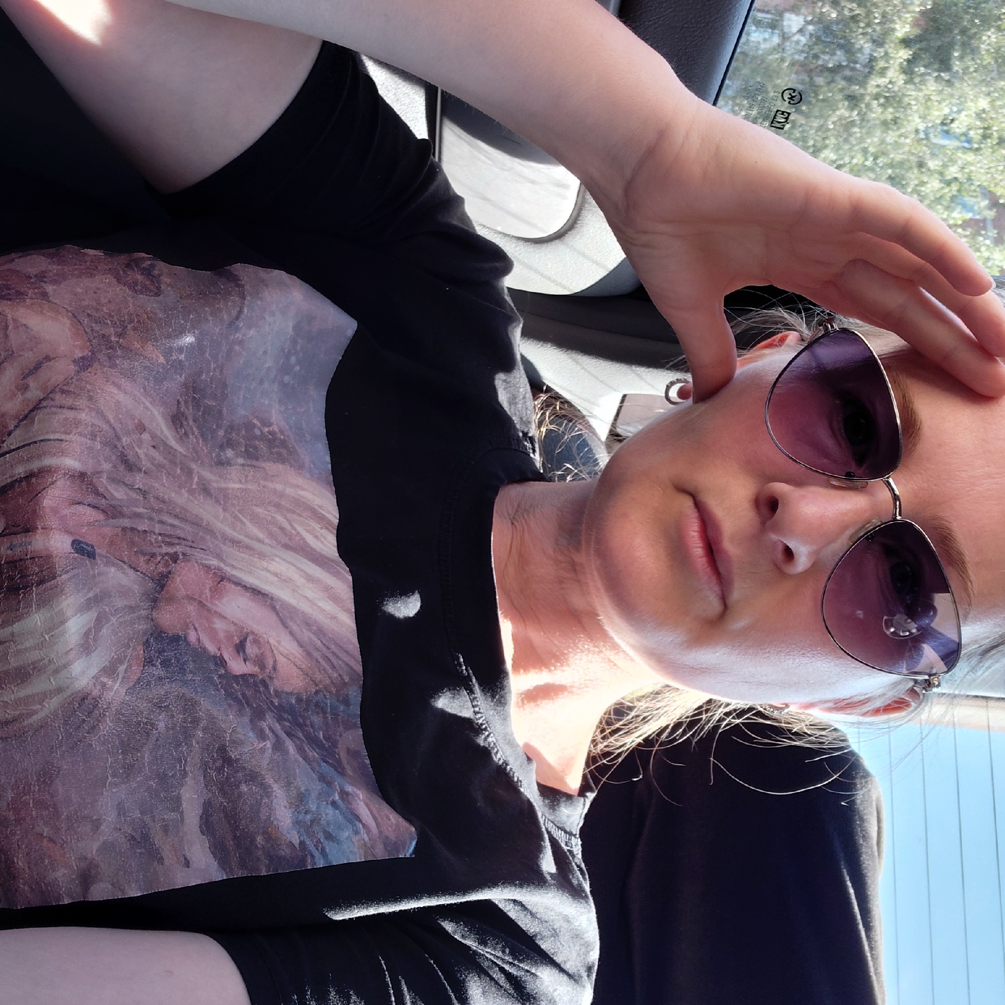 A woman with blonde hair, wearing sunglasses and a black t-shirt with a portrait of a woman on it, is sitting in the backseat of a car. Her hand is on her forehead. Sunlight streams in from the side. The background shows trees and a blue sky.