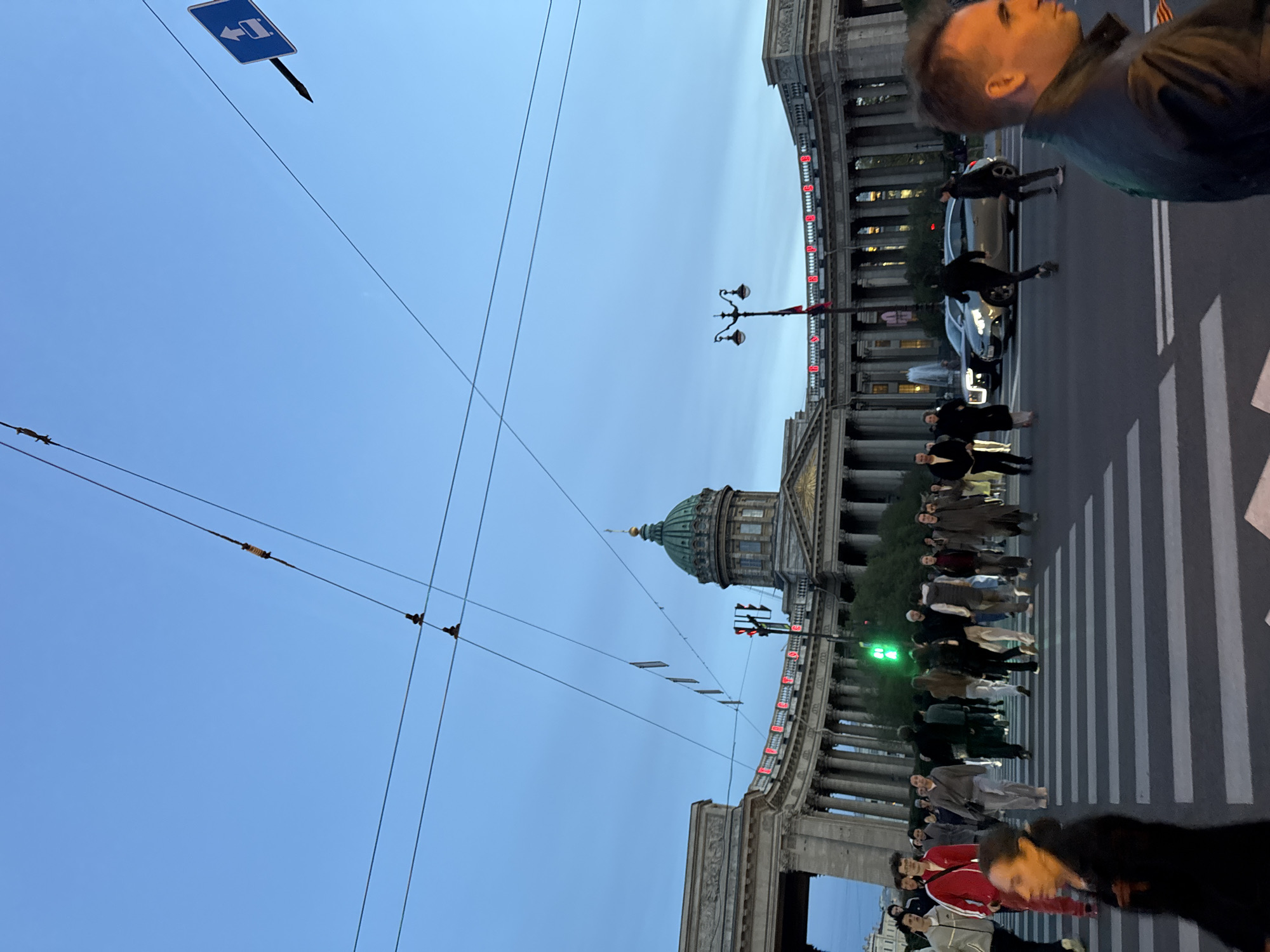The image shows a street scene in Saint Petersburg, Russia, at dusk. The Kazan Cathedral is visible with its green dome. A crowd of people is crossing the street at a pedestrian crosswalk. A car is stopped at the crosswalk. A man is visible in the foreground, facing away from the camera. Overhead, power lines crisscross the sky, and a blue street sign is mounted on a pole. A row of buildings with columns lines the street. The lights on the building are red. A street lamp is near the cathedral. A man can be seen walking behind the car.