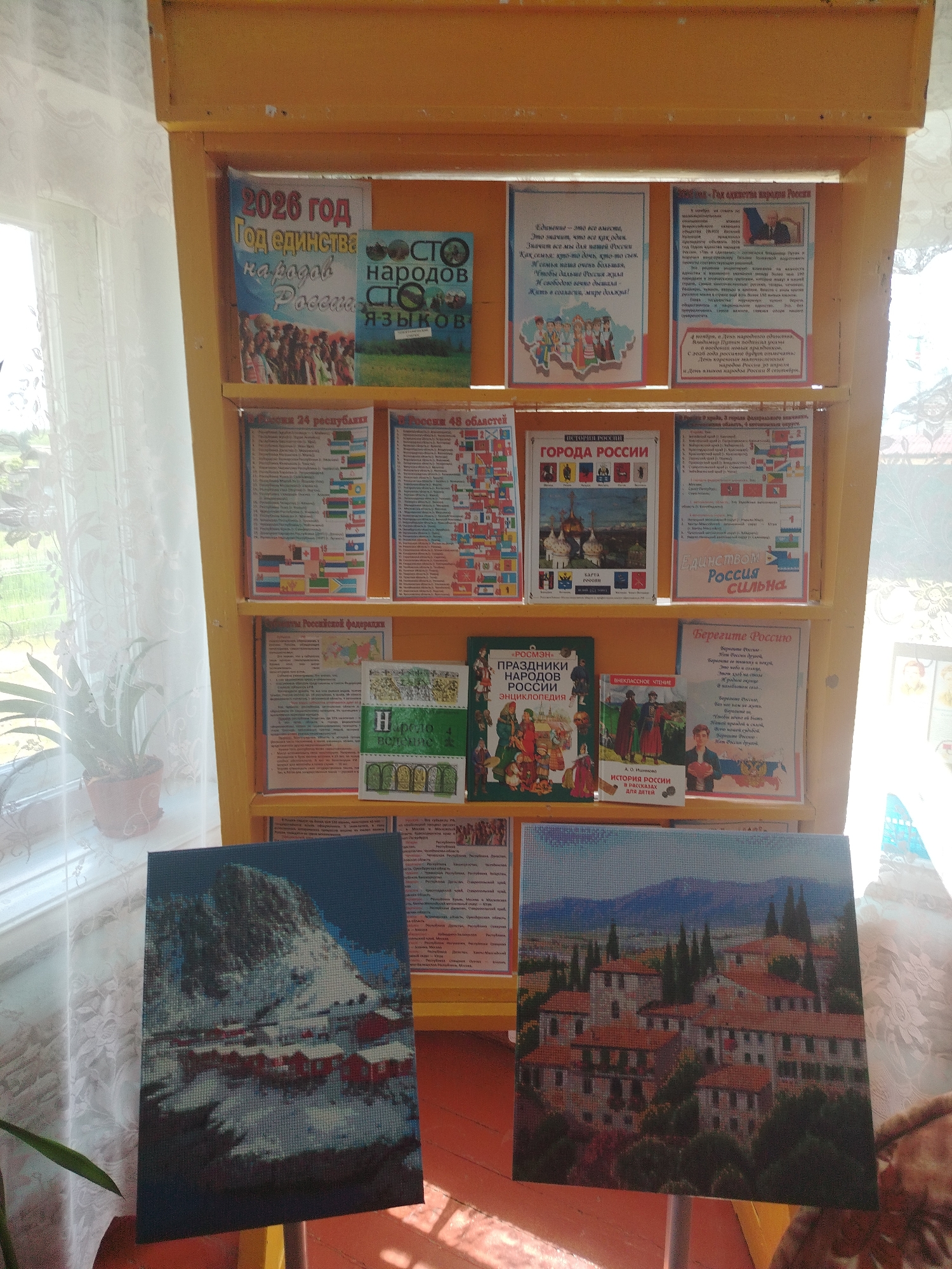 The image displays a yellow shelf unit filled with various posters, books, and two framed diamond paintings. The posters are predominantly themed around Russian history, culture, and unity. One poster is titled "2026 год Год единства народов России" (2026 Year Year of Unity of Peoples of Russia). Another poster features an illustration of various ethnic groups and is labeled "Сто народов Сто языков" (One Hundred Peoples, One Hundred Languages). Several other posters detail Russian regions, cities, and historical facts. There are also books titled "Праздники народов России. Энциклопедия" (Holidays of Peoples of Russia. Encyclopedia) and "История России. Рассказы для детей" (History of Russia. Stories for Children).
The two diamond paintings at the bottom depict landscapes. The left painting shows a snow-covered mountain village with red houses, while the right painting showcases an Italian-style town with terracotta roofs and cypress trees. The shelf unit is positioned indoors, near a window with white curtains, suggesting it's in a room within a building. The overall scene appears to be a display or a cultural exhibit. The specific location within the city of Klyuchi, Russia, cannot be determined from the image itself. The background suggests a domestic or educational setting. There are no people visible in the image.
